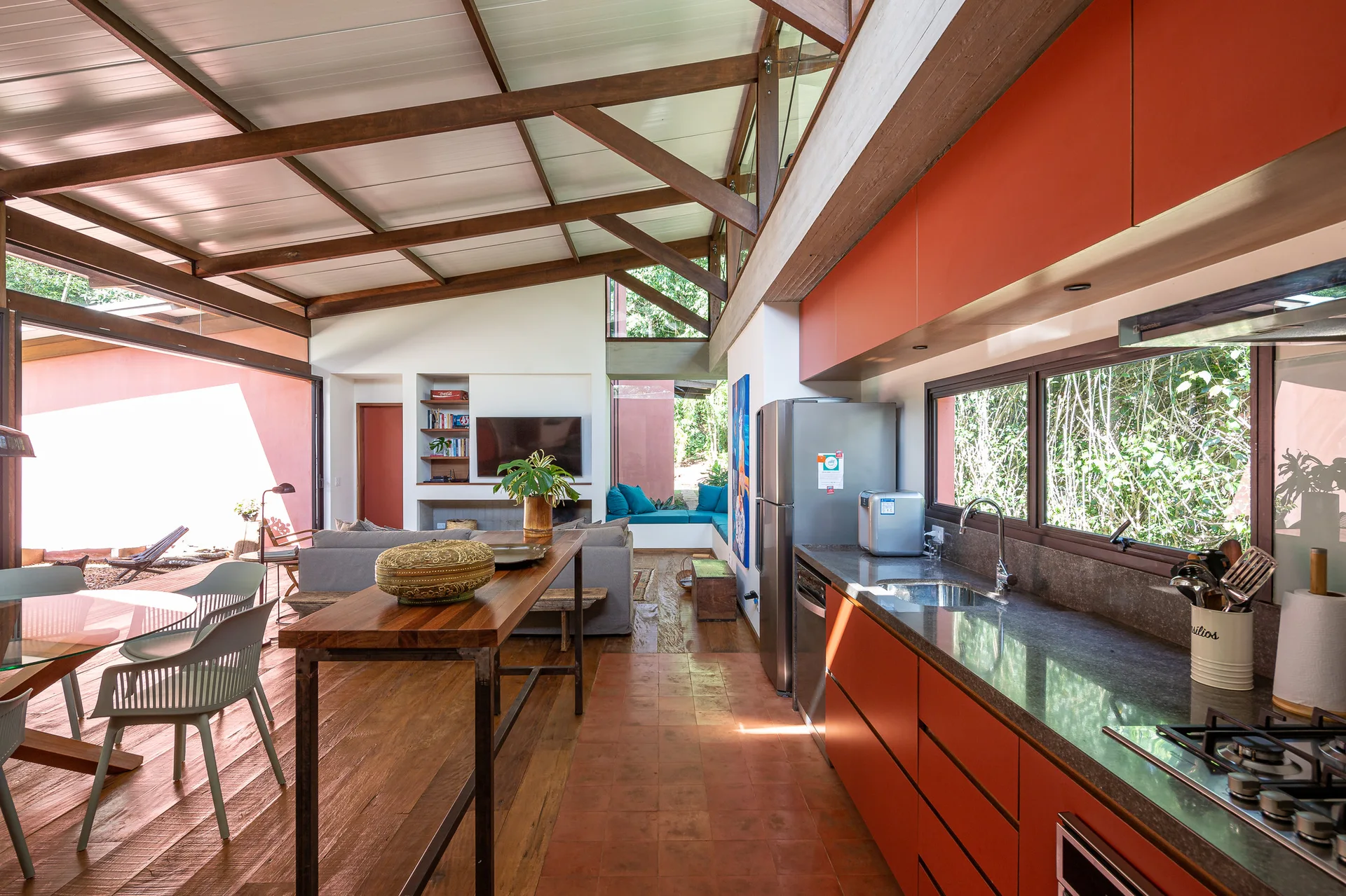 An open-concept kitchen features vibrant orange cabinets, a stainless steel refrigerator, dark wood ceiling beams, and a wooden console table.