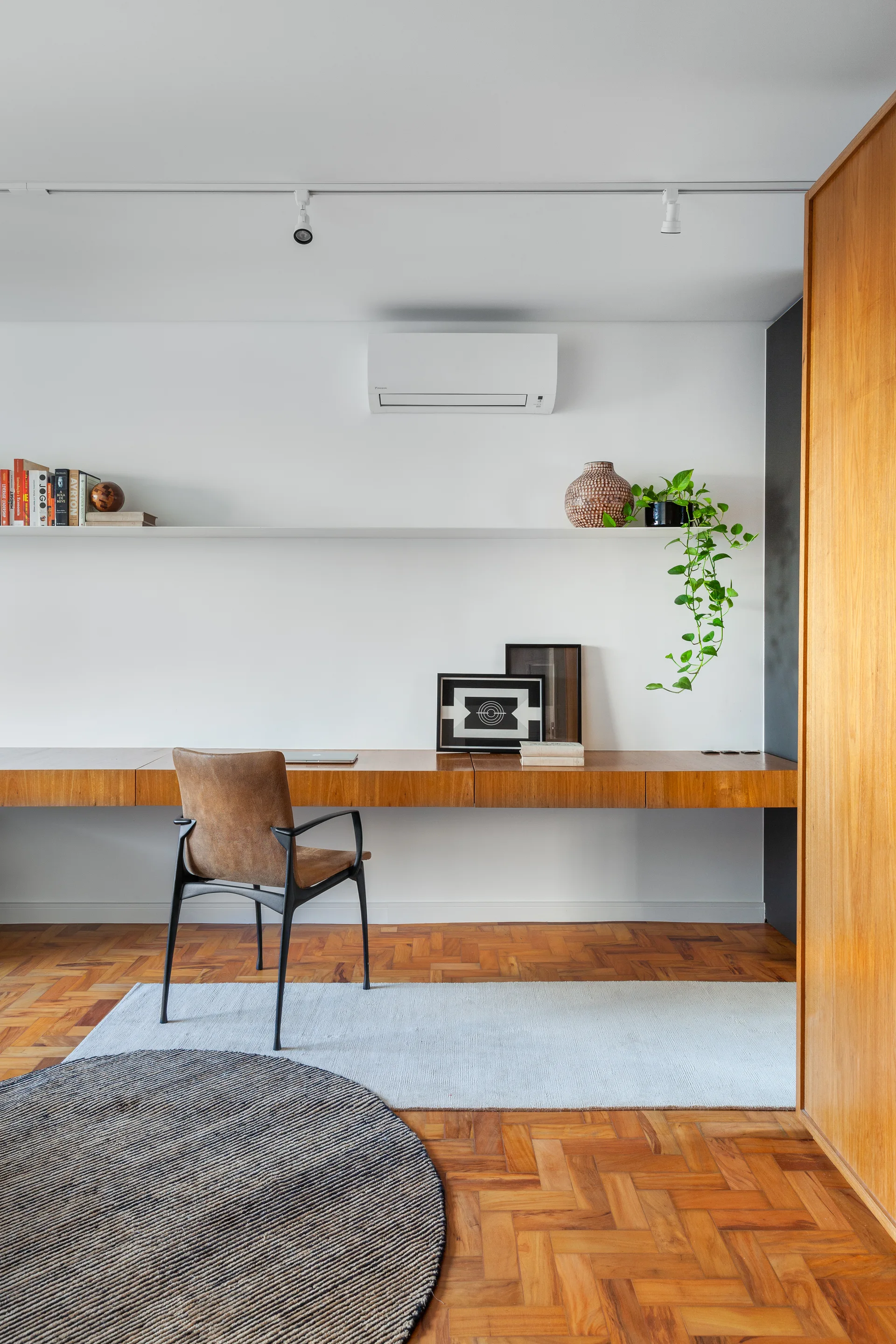 A minimalist home office featuring a long floating wooden desk, a brown leather chair, a floating white shelf, and parquet wood flooring.