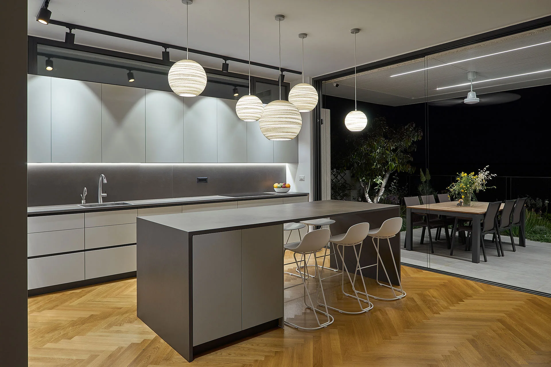 A modern grey kitchen features a central island with white bar stools, herringbone wood floors, and sliding glass doors opening to an outdoor patio.