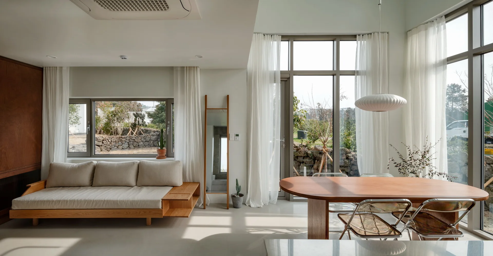 A minimalist living room features a wooden platform sofa, an oval dining table with clear acrylic chairs, and large windows with sheer white curtains.