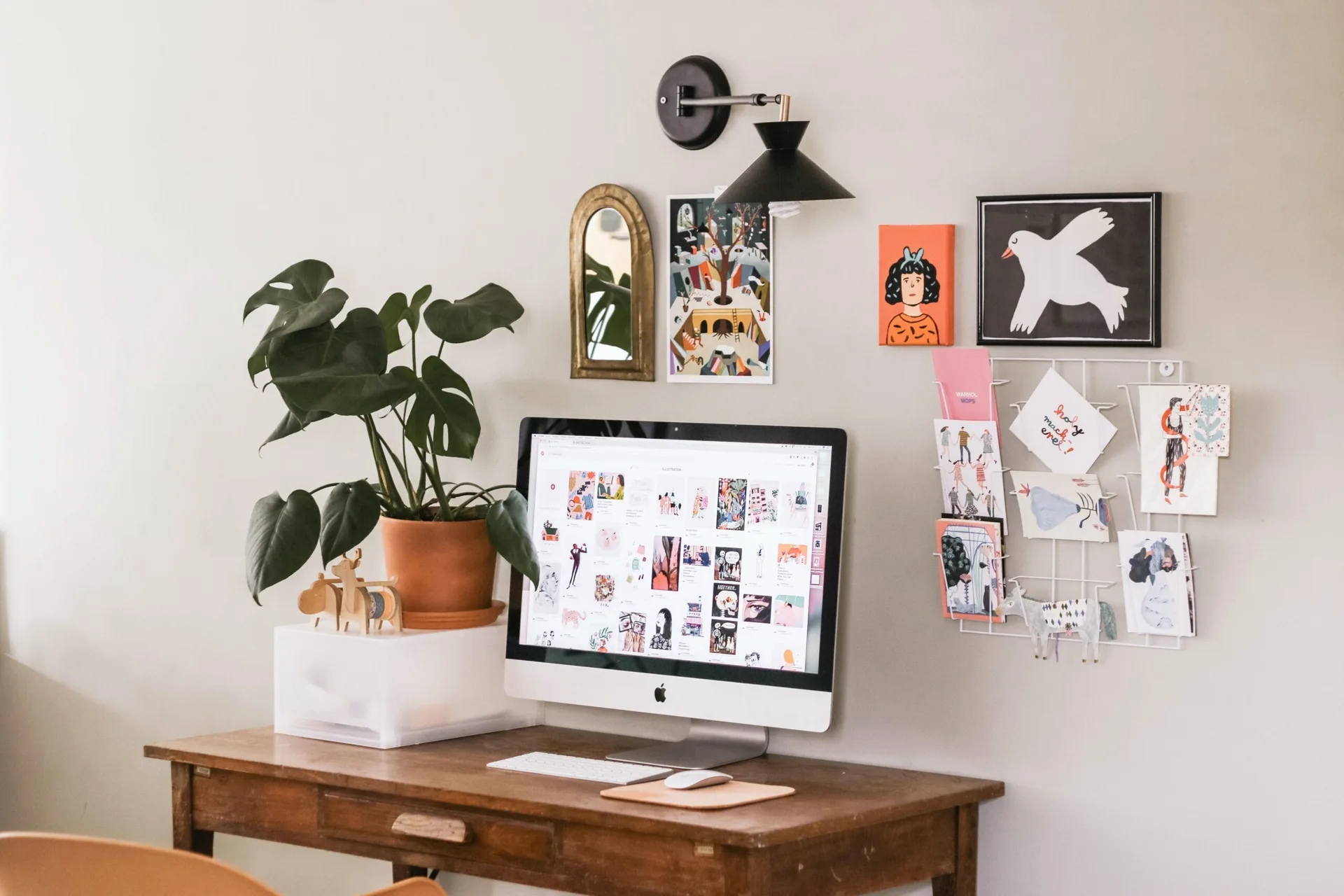 A desktop computer sits on a vintage wooden desk below a black sconce, flanked by a potted monstera plant and an eclectic gallery wall of art prints.