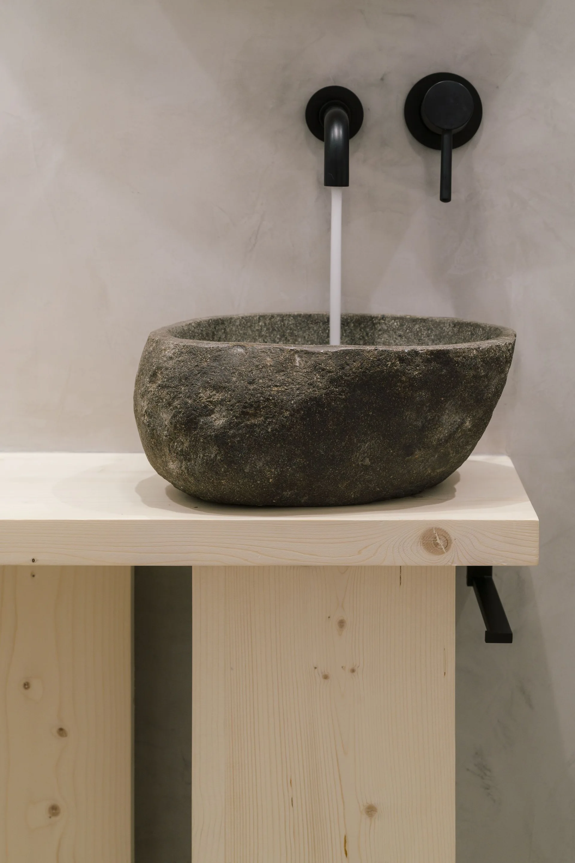 A rounded grey stone vessel sink sits on a light-colored natural wood vanity with a wall-mounted matte black faucet pouring water.