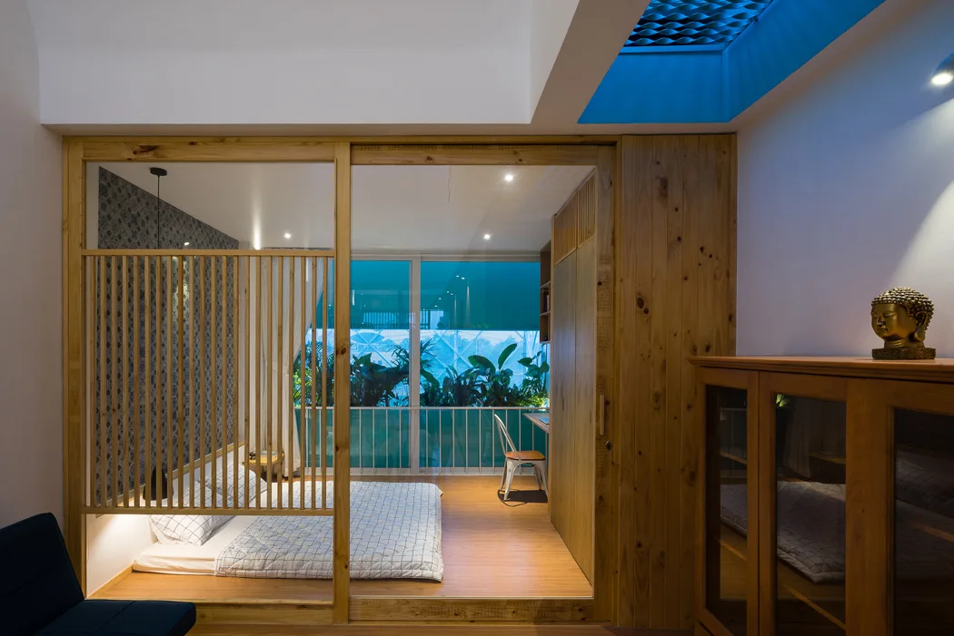 A minimalist bedroom features a floor mattress behind a wooden slatted sliding partition next to a large window and a blue-lit skylight.