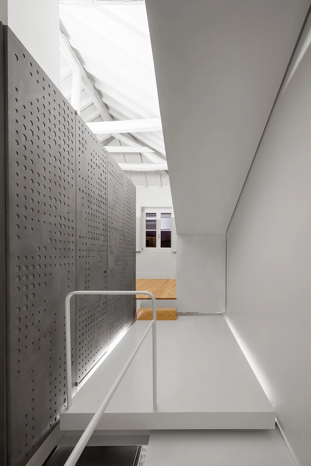 A minimalist white hallway featuring a large perforated grey metal partition wall, stairs, and exposed ceiling beams.