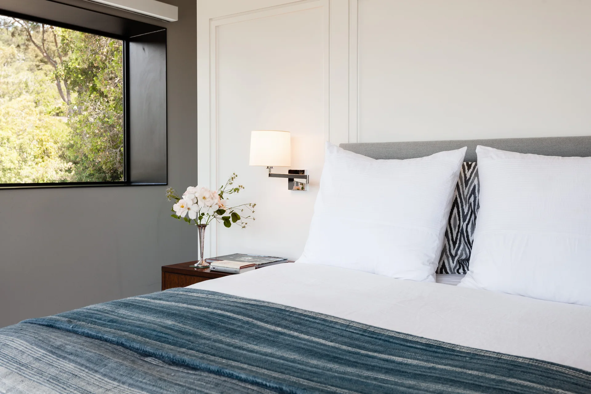 A minimalist bedroom features a grey bed with blue linens, a wooden nightstand, a wall-mounted sconce, and a window overlooking green trees.