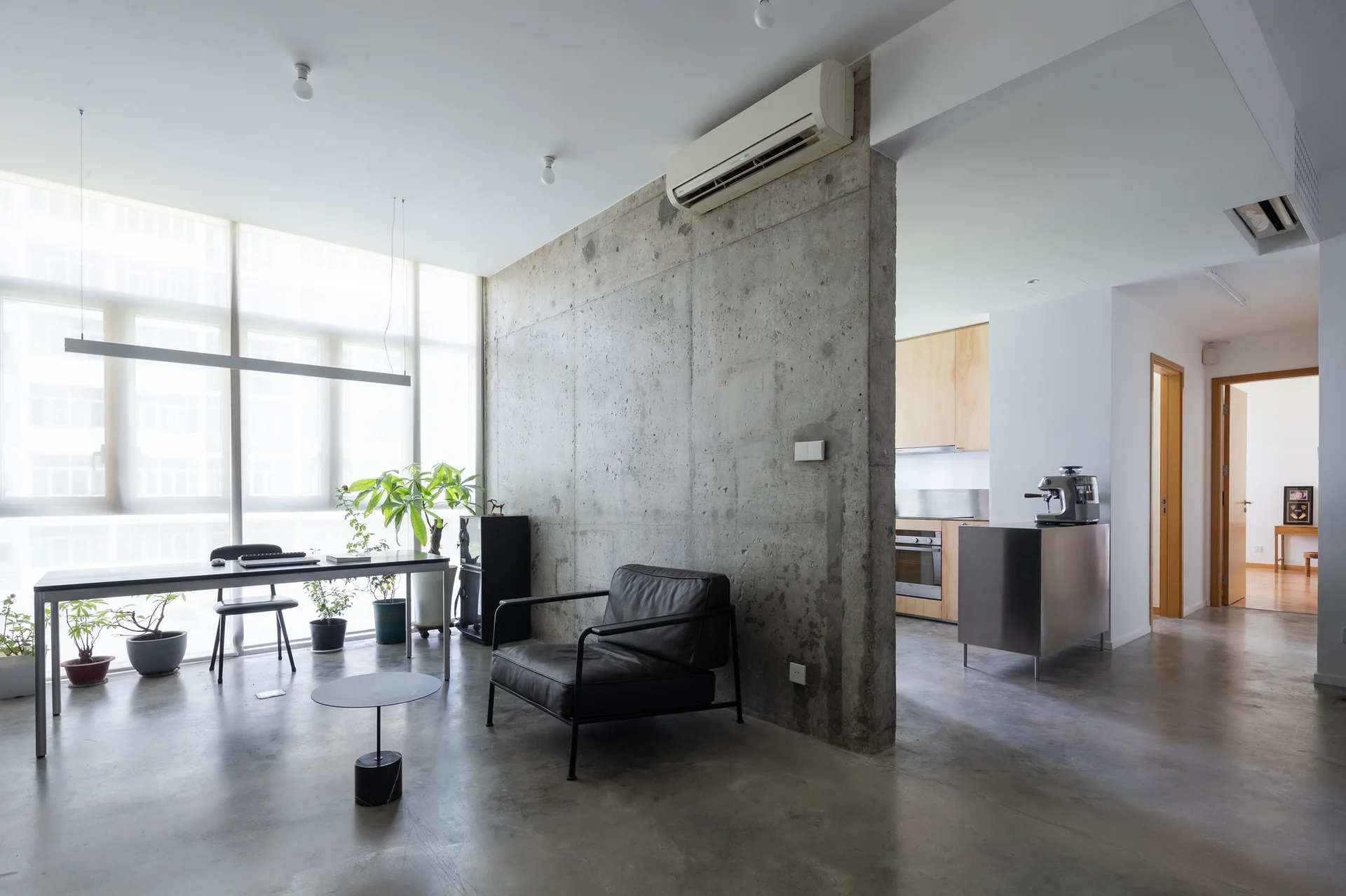 A minimalist living space with an exposed concrete partition wall, a black leather armchair, and a slim desk next to a bright window.