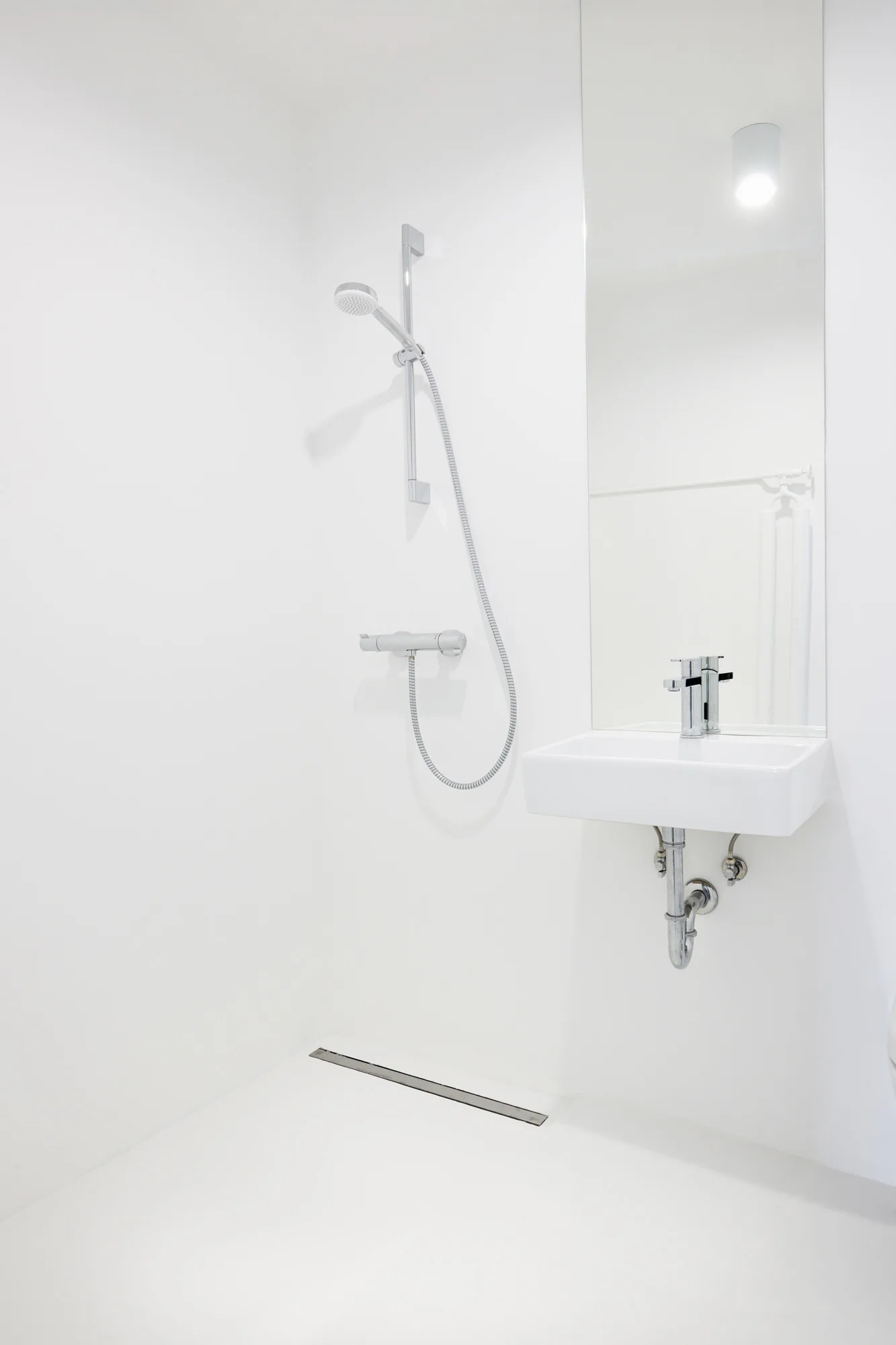 A minimalist all-white bathroom with a wall-mounted sink, a tall frameless mirror, and a chrome shower set.