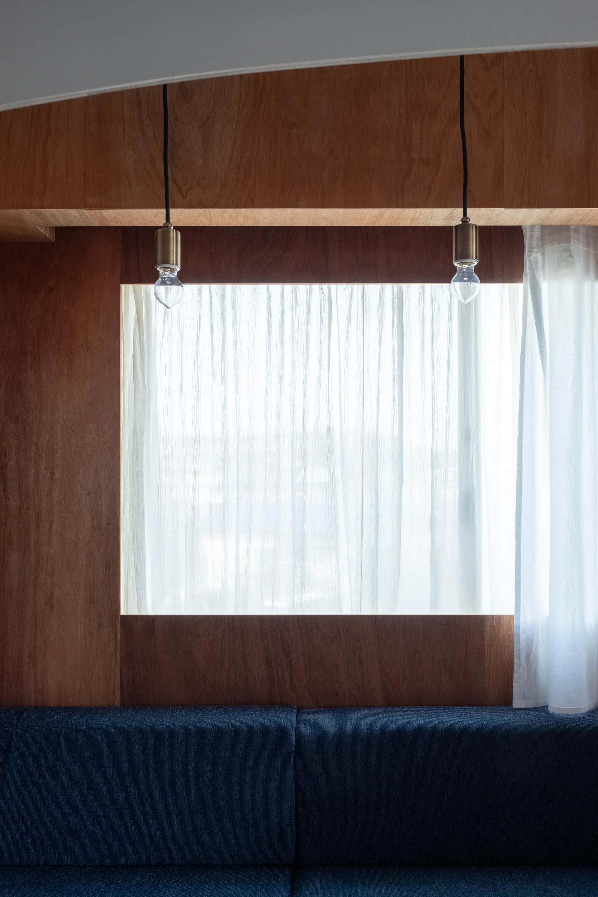 A dark blue sofa sits against a wood-paneled wall under an inset window with sheer white curtains and hanging bare-bulb pendant lights.