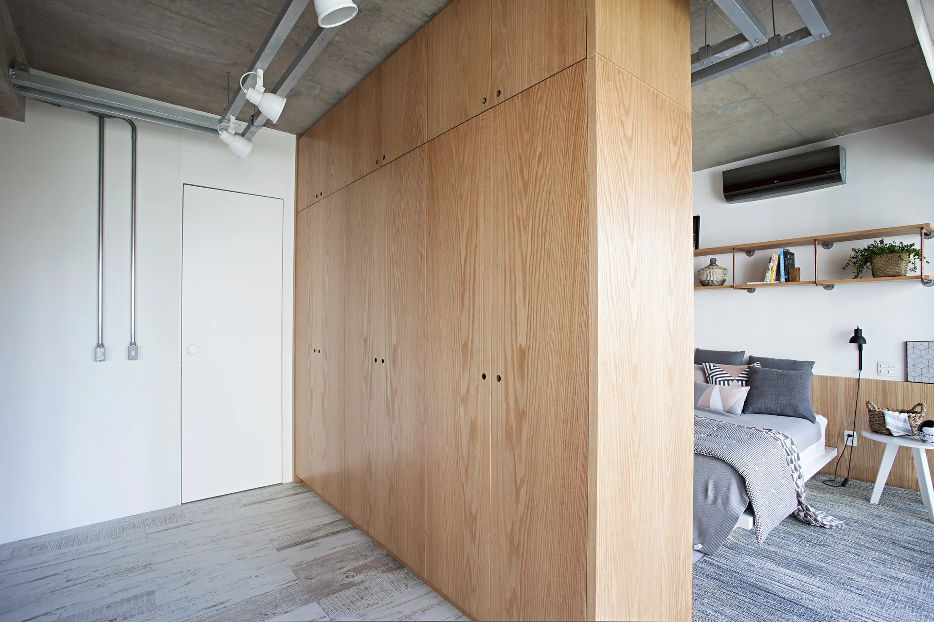 A light wood floor-to-ceiling wardrobe partition stands next to a bed with grey linens beneath an exposed concrete ceiling with track lights.
