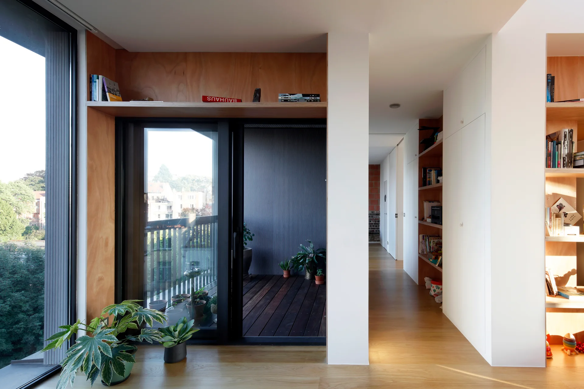 A modern residential room featuring custom plywood built-in shelving, a sliding glass door to a balcony, and white walls.