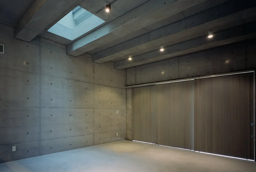 A minimalist raw concrete room features structural ceiling beams, a rectangular skylight, spotlight fixtures, and large wood sliding panels.