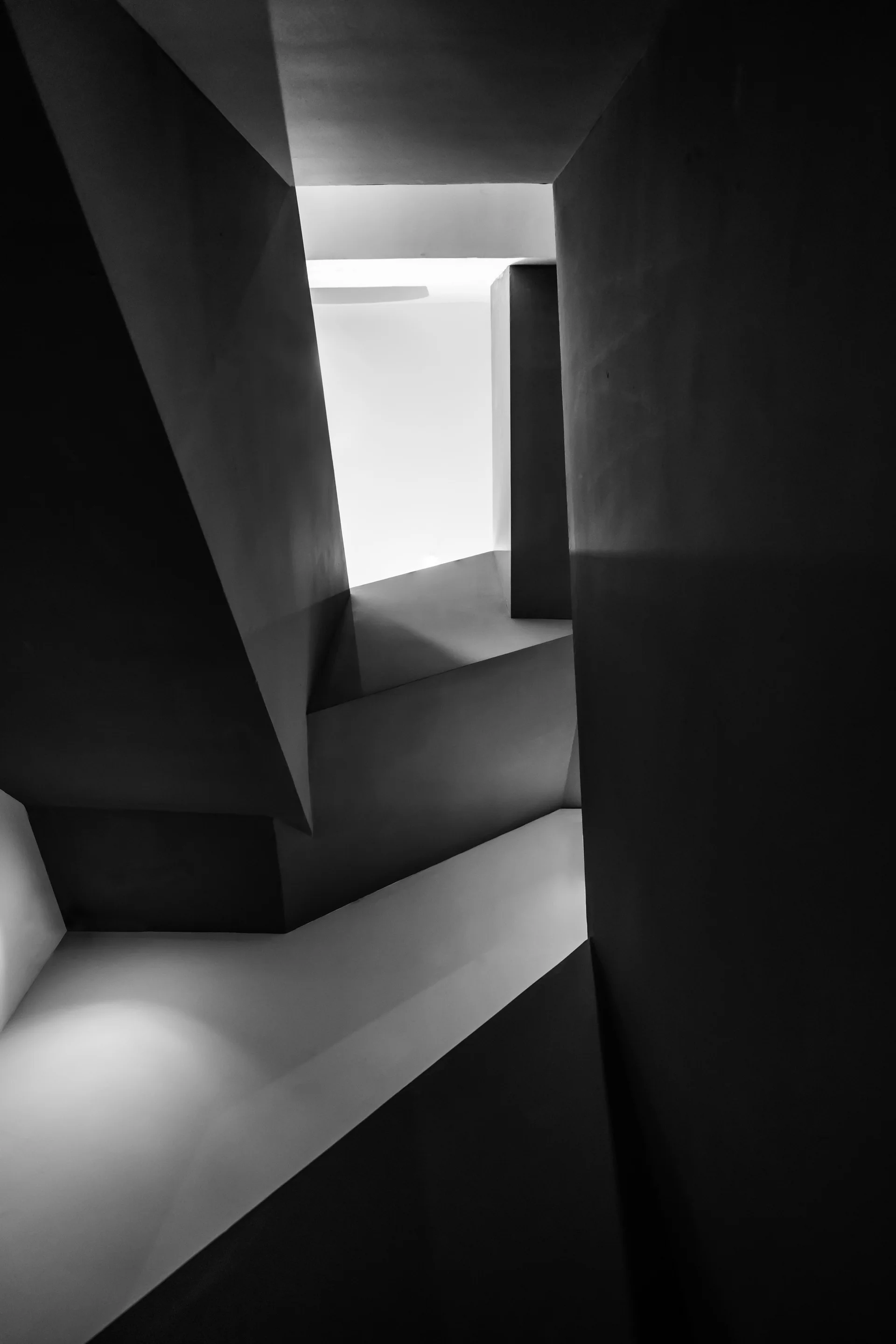 An abstract black and white photograph of geometric concrete walls intersecting to create sharp angles in a stairwell.