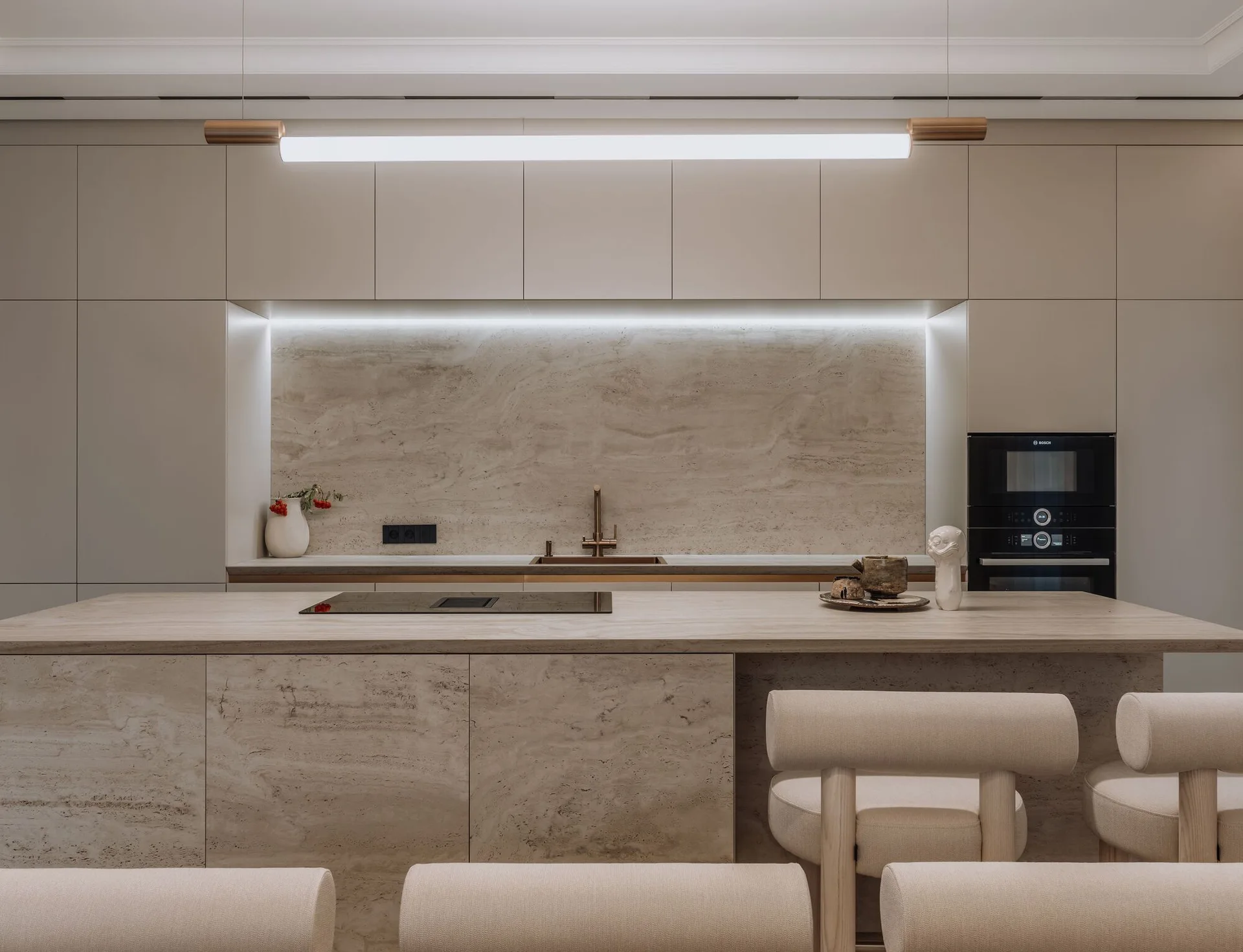 A minimalist kitchen featuring a stone-faced island with stools, a matching stone backsplash, and neutral handleless cabinetry.