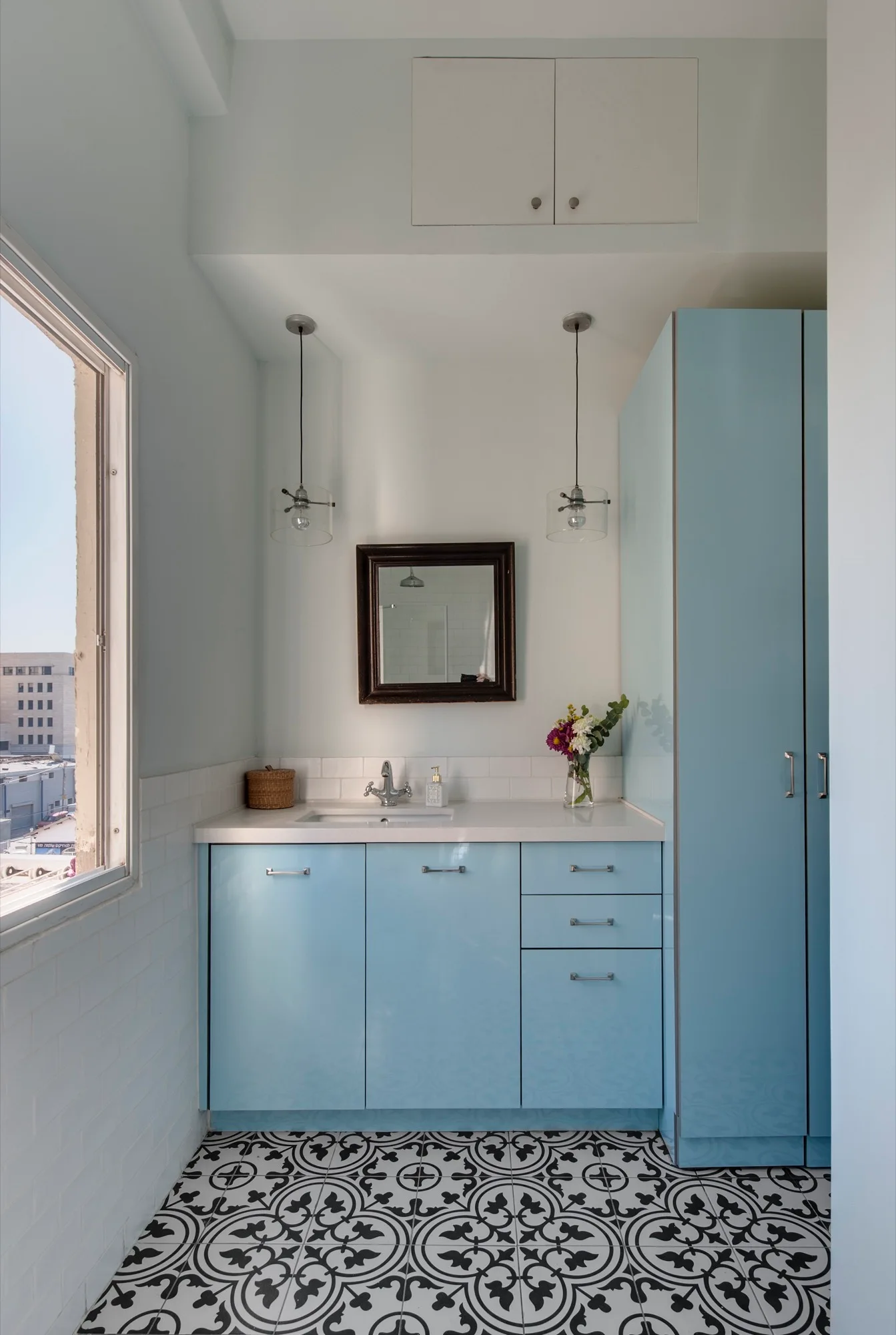 A light blue bathroom vanity and matching tall cabinet stand on a patterned black and white tile floor next to a window.