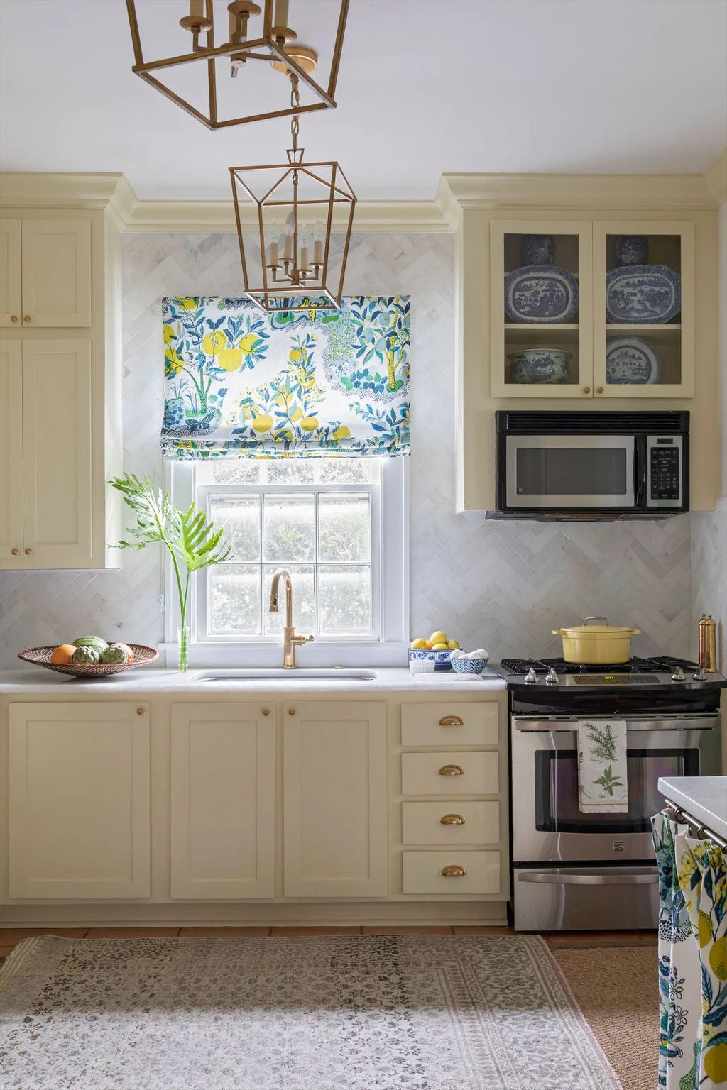 A cream kitchen featuring a herringbone stone backsplash, a sink with a brass faucet, and a window dressed with a botanical Roman shade.