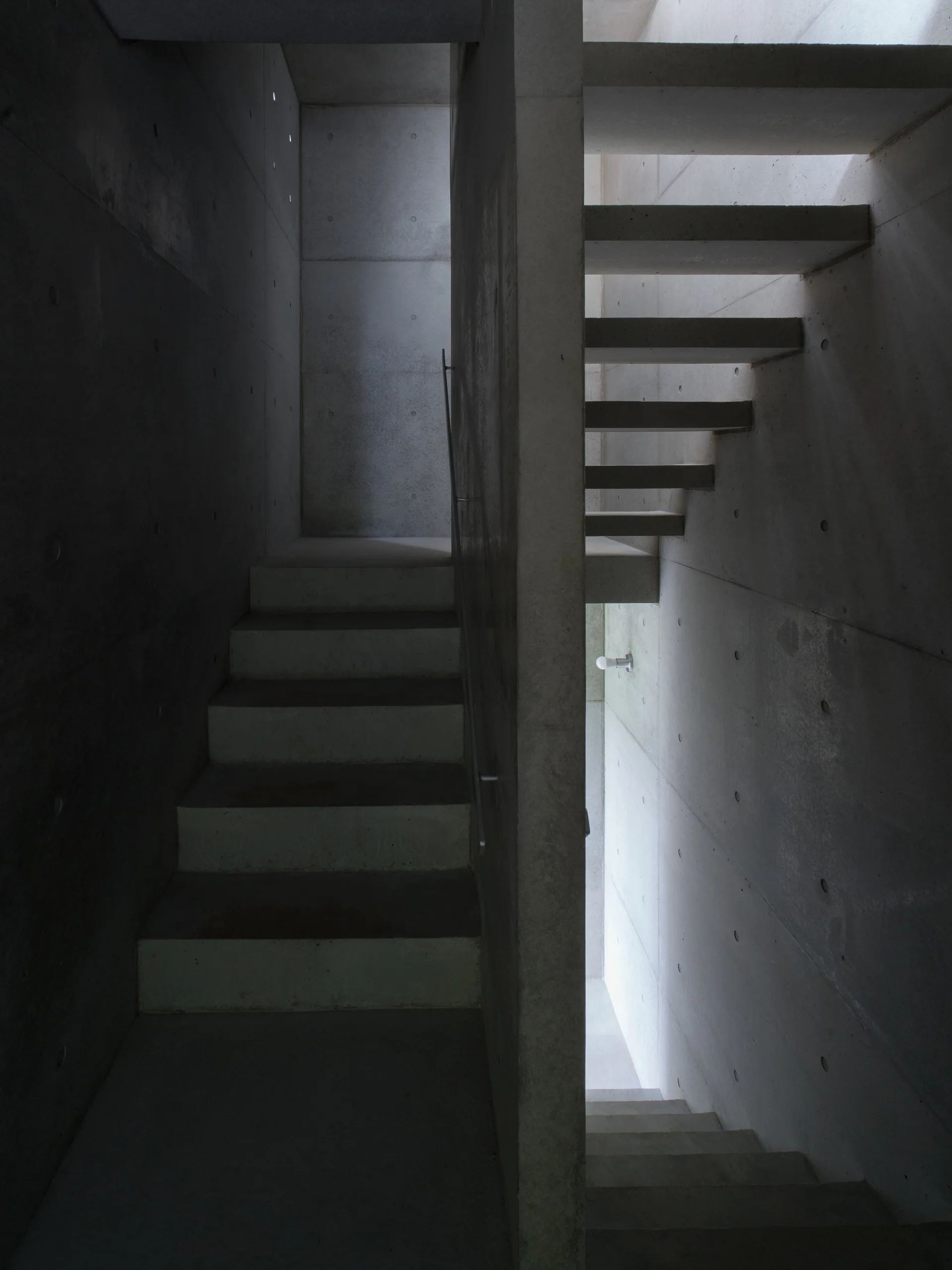 A minimalist cast concrete stairwell features floating concrete treads and smooth grey walls with uniform tie rod holes under diffused natural light.
