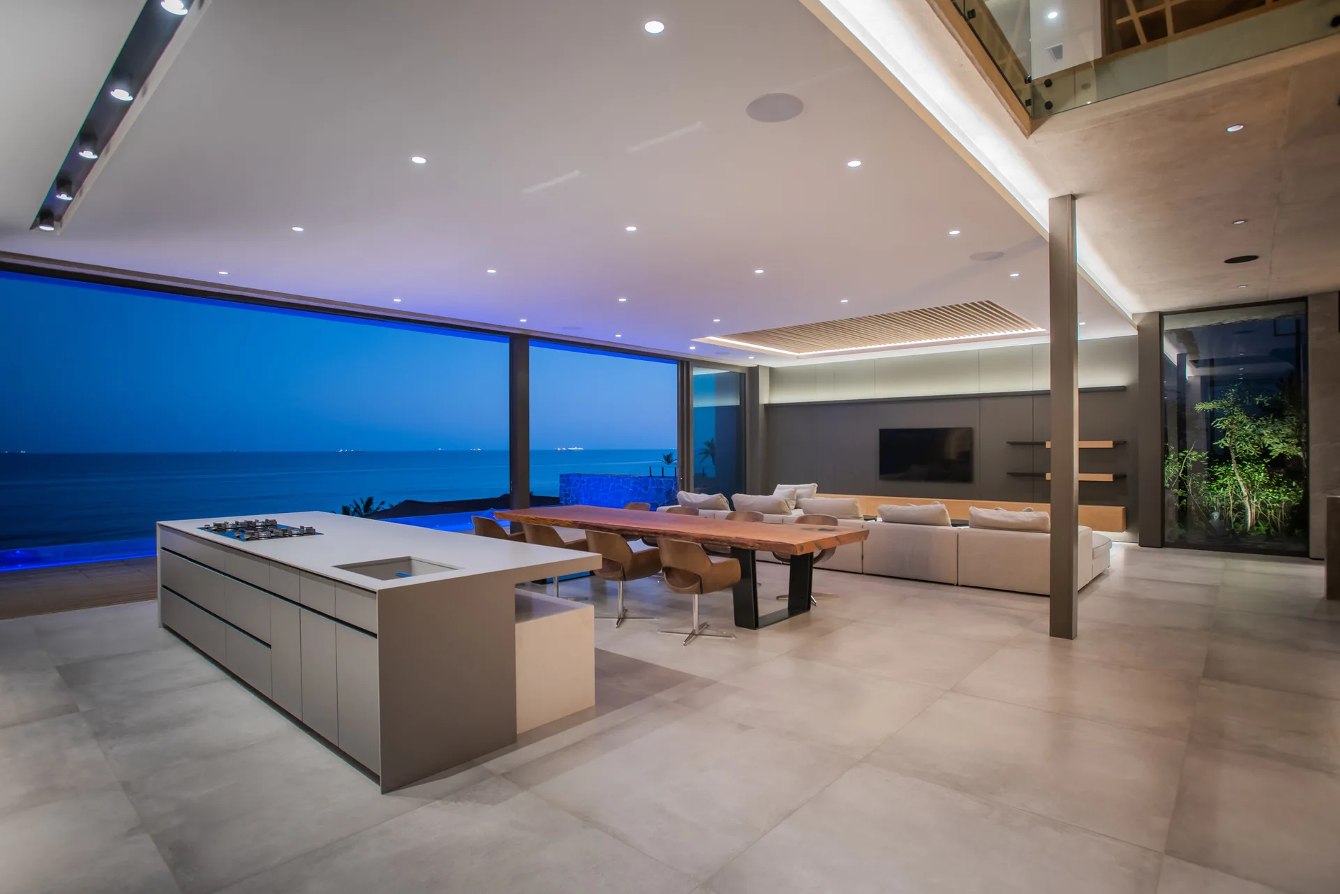 An open-concept living space features a grey kitchen island, a long live-edge wooden dining table, and large glass walls overlooking the ocean.