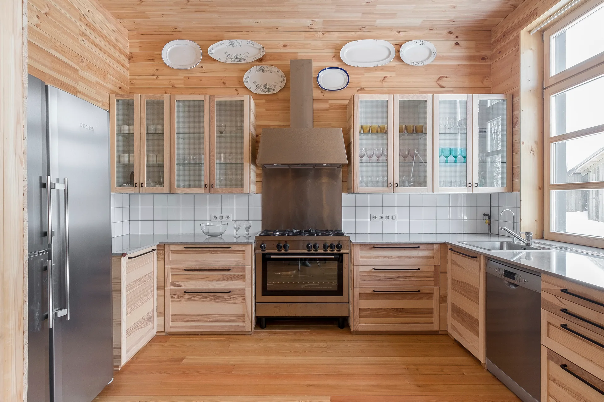A kitchen featuring horizontal pine wood walls, matching wooden cabinetry with glass doors, a white square tile backsplash, and stainless steel appliances.