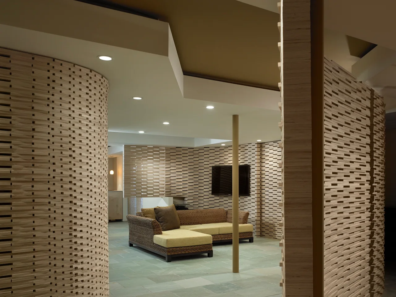 A living room features curved wooden slat partition walls framing a wicker sectional sofa on green stone tile flooring.