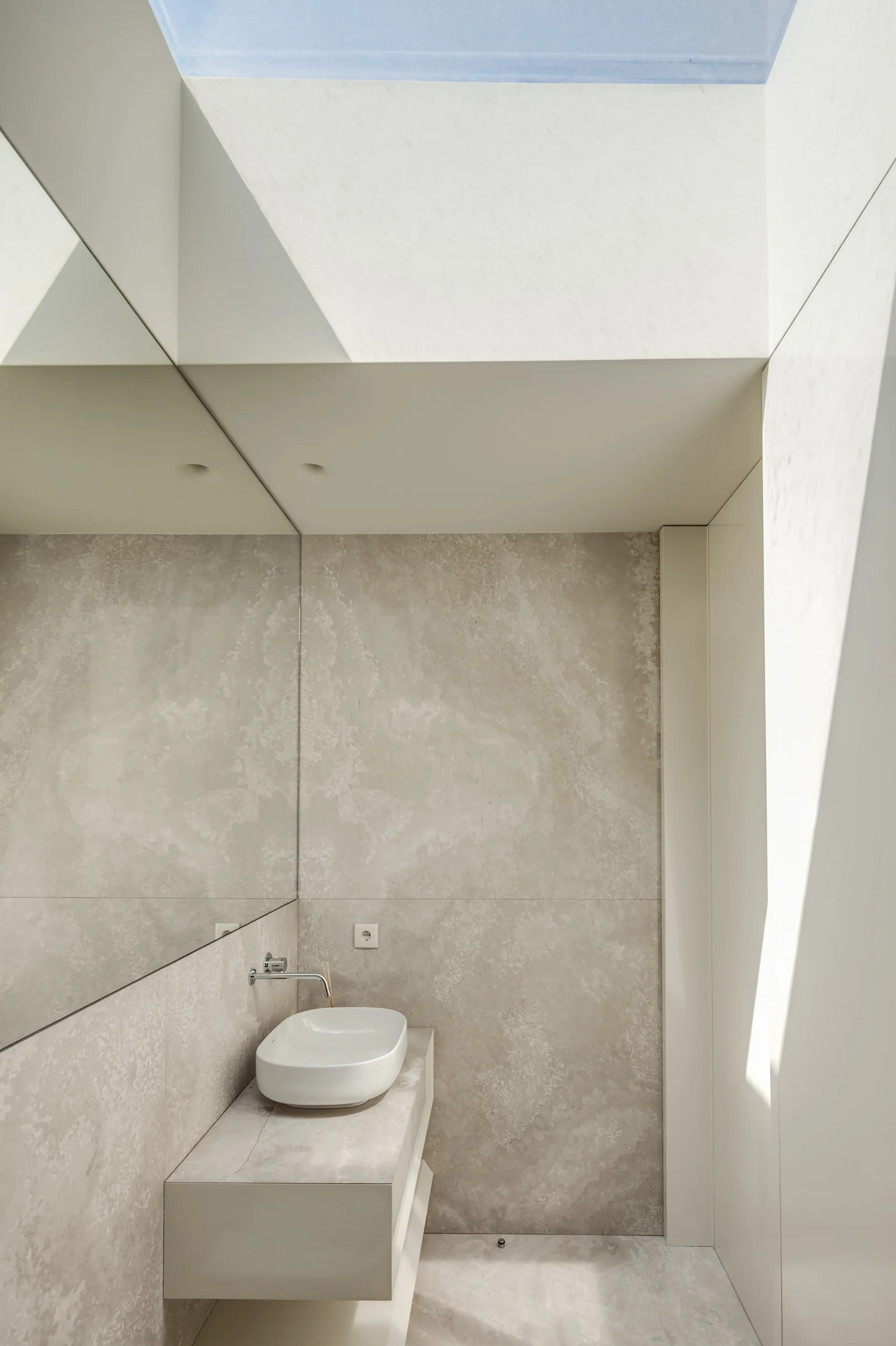 A minimalist bathroom features a floating beige stone vanity with a white vessel sink, a large wall mirror, and natural light from a skylight above.
