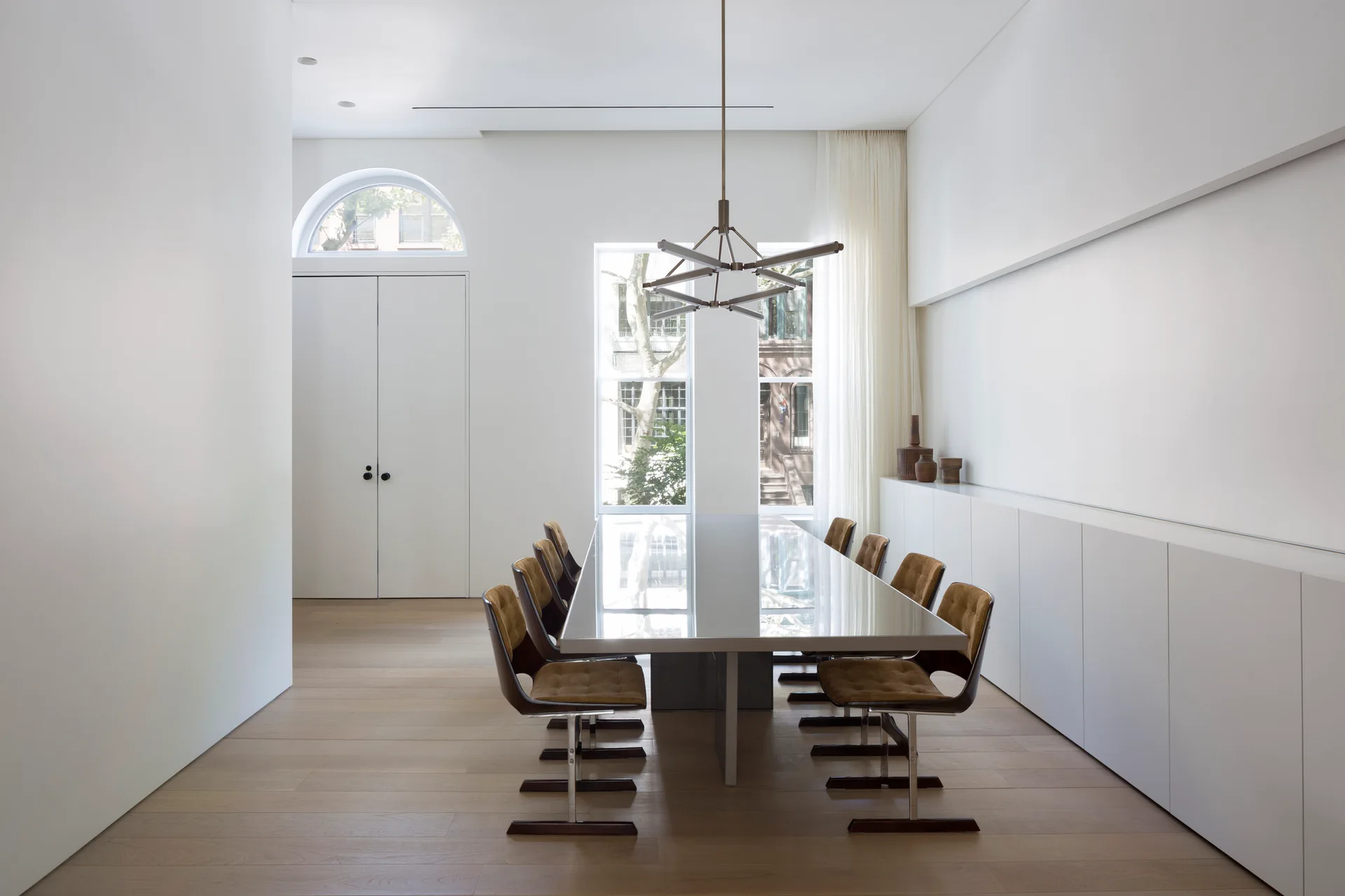 A minimalist dining room features a long glossy gray table surrounded by eight brown upholstered chairs, set against white walls and light wood flooring.