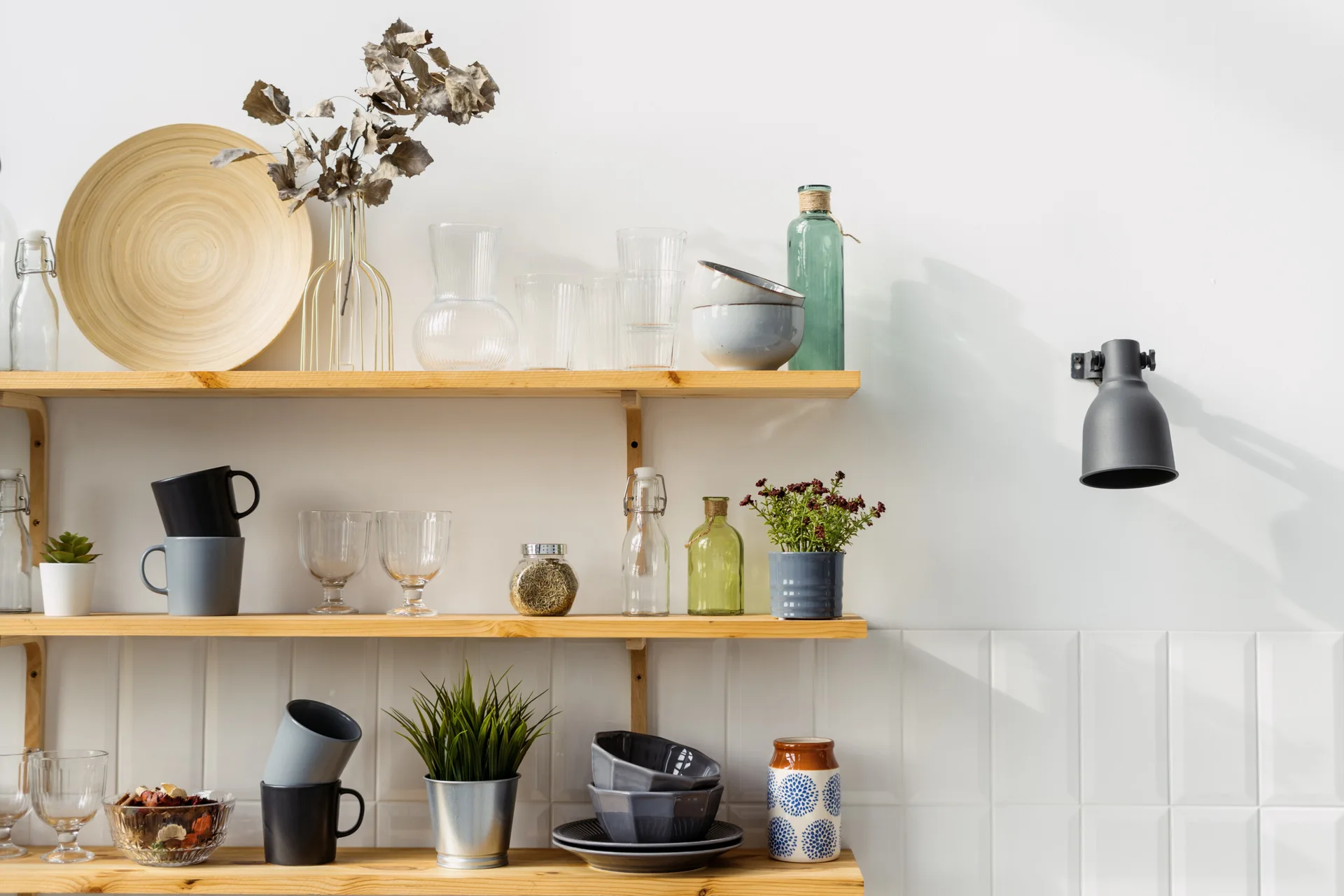Three-tier light wood open shelves display mugs, bowls, and glassware against a white tiled kitchen wall next to a black wall-mounted lamp.