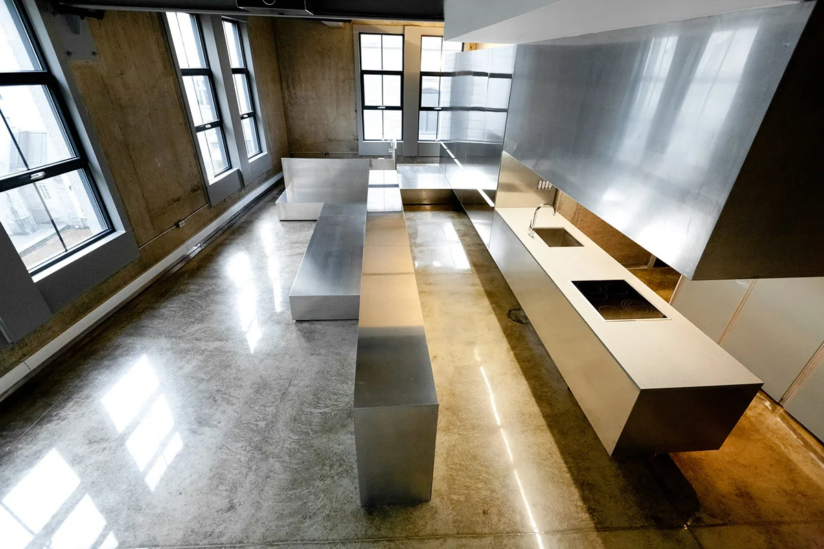 An industrial loft kitchen features extensive stainless steel cabinetry and kitchen islands set against raw concrete walls and polished floors.
