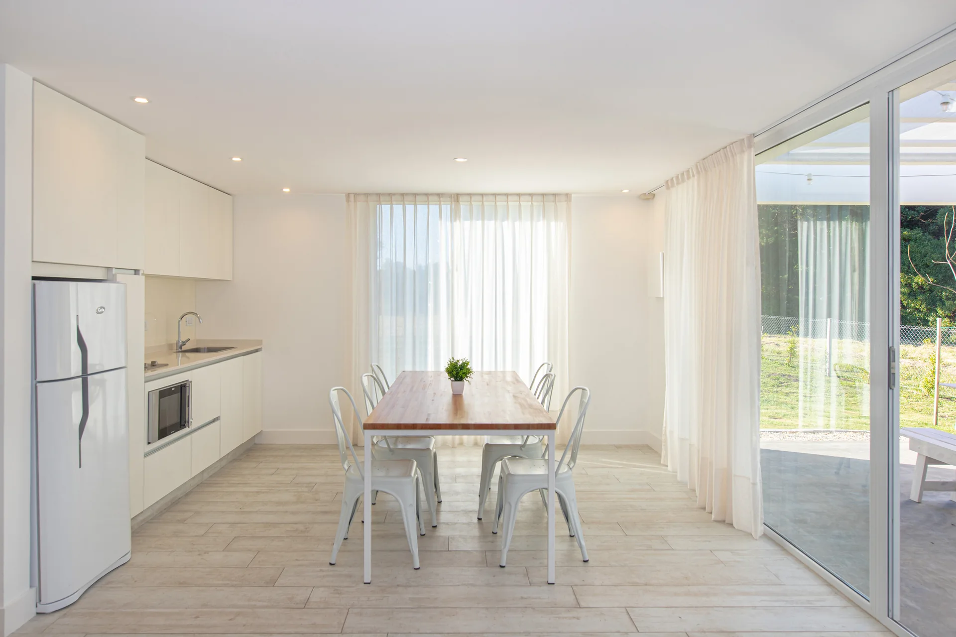 A bright kitchen and dining room featuring white minimalist cabinets, a wood-topped dining table with white metal chairs, and sheer floor-length curtains.