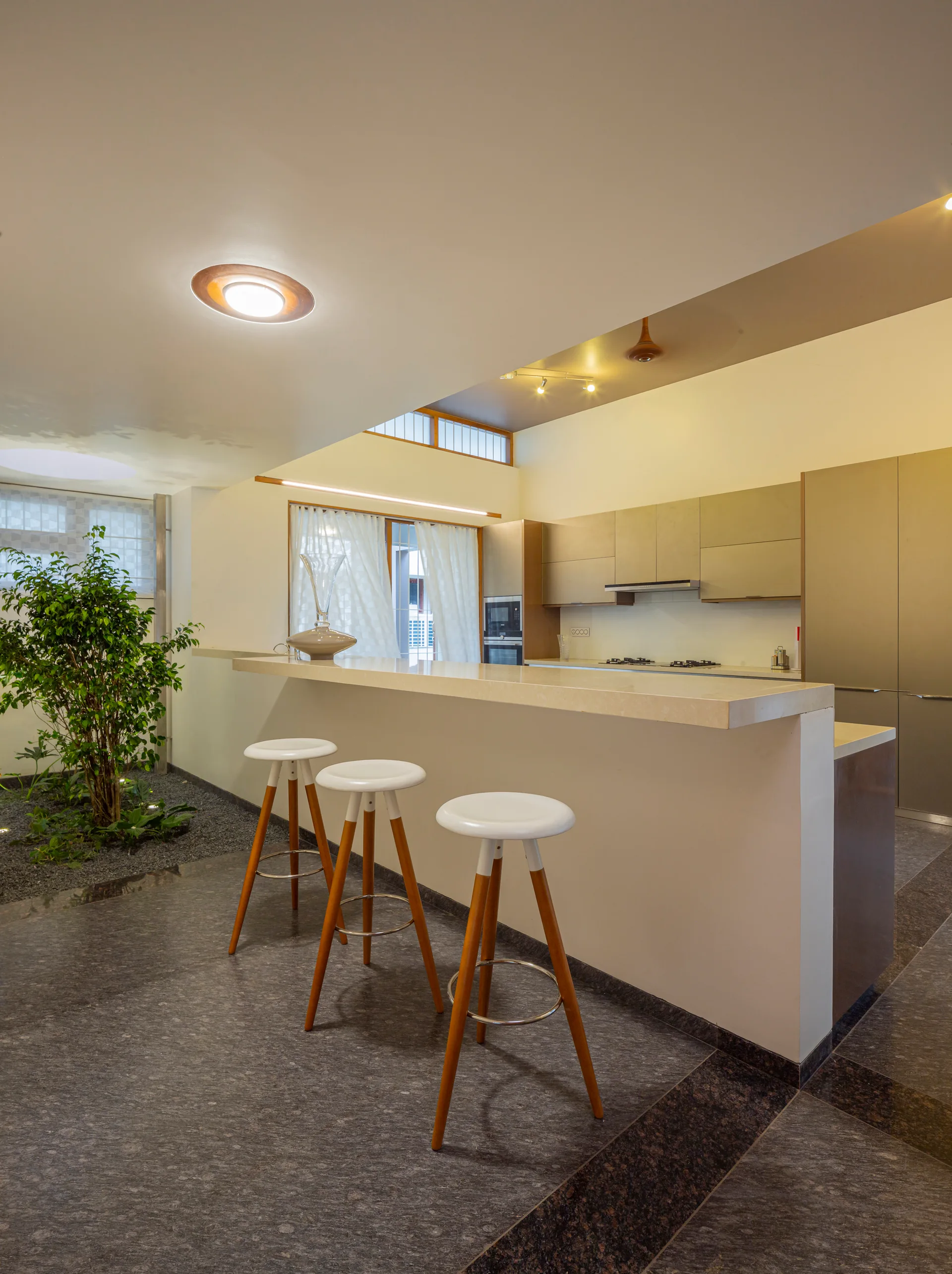 A minimalist kitchen featuring a beige breakfast bar with three wooden stools next to an indoor planting bed with dark gravel.
