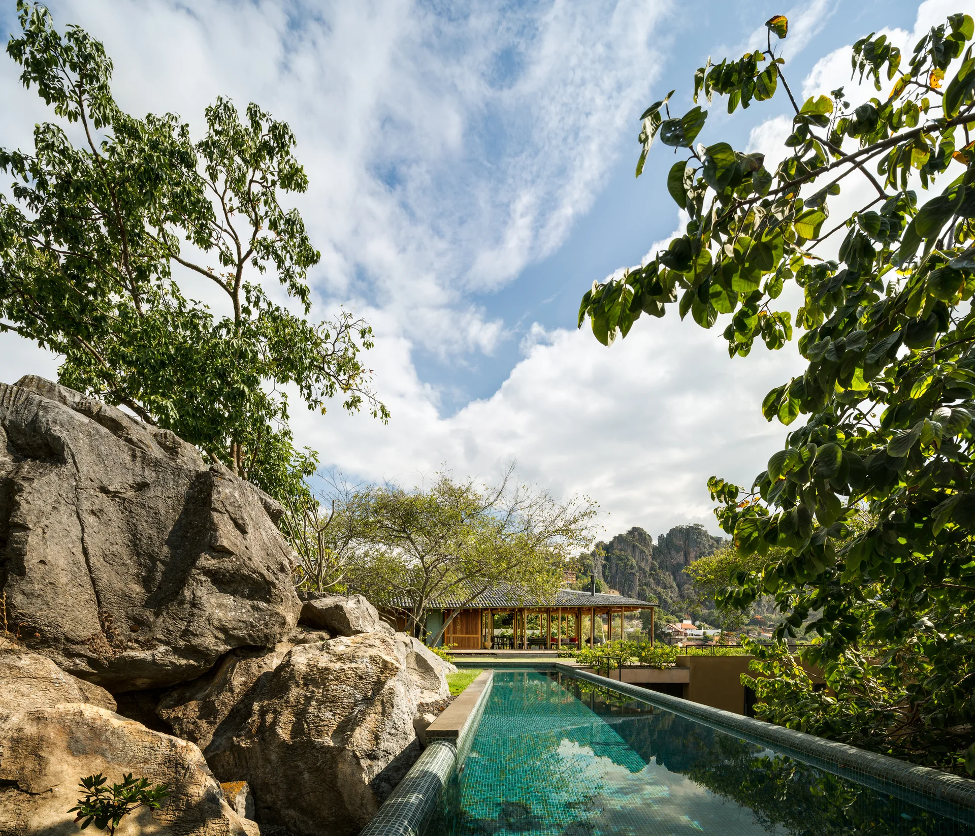 A long turquoise mosaic tiled lap pool runs alongside massive gray rock boulders with a wooden pavilion and mountains in the distance.