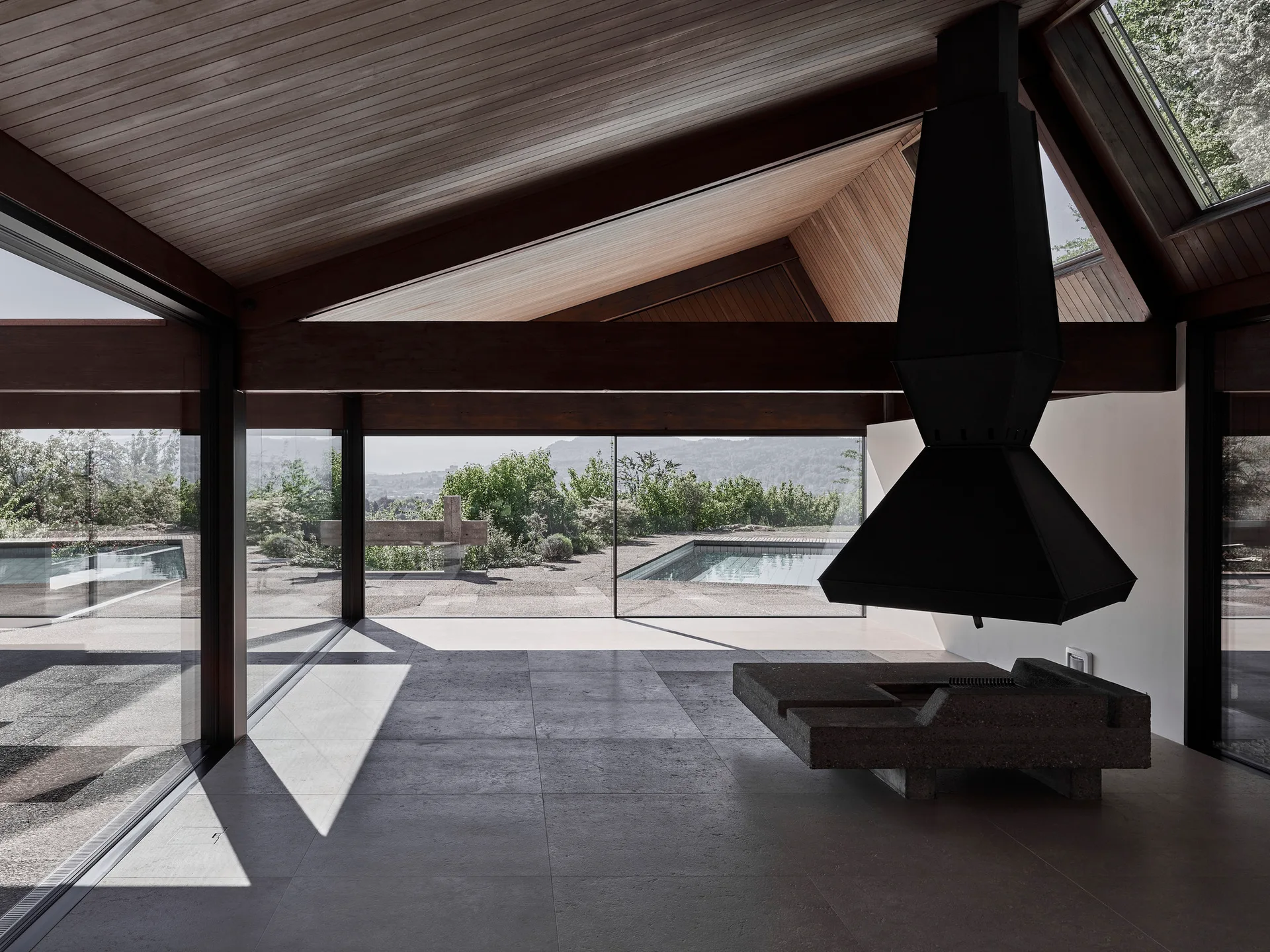 A minimalist room features a suspended black metal fireplace above a stone hearth, a sloped wood ceiling, and floor-to-ceiling glass walls.