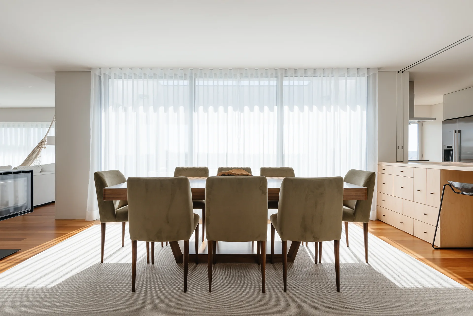 A modern dining room features a rectangular wooden table and velvet-upholstered chairs set against a large window with sheer white curtains.