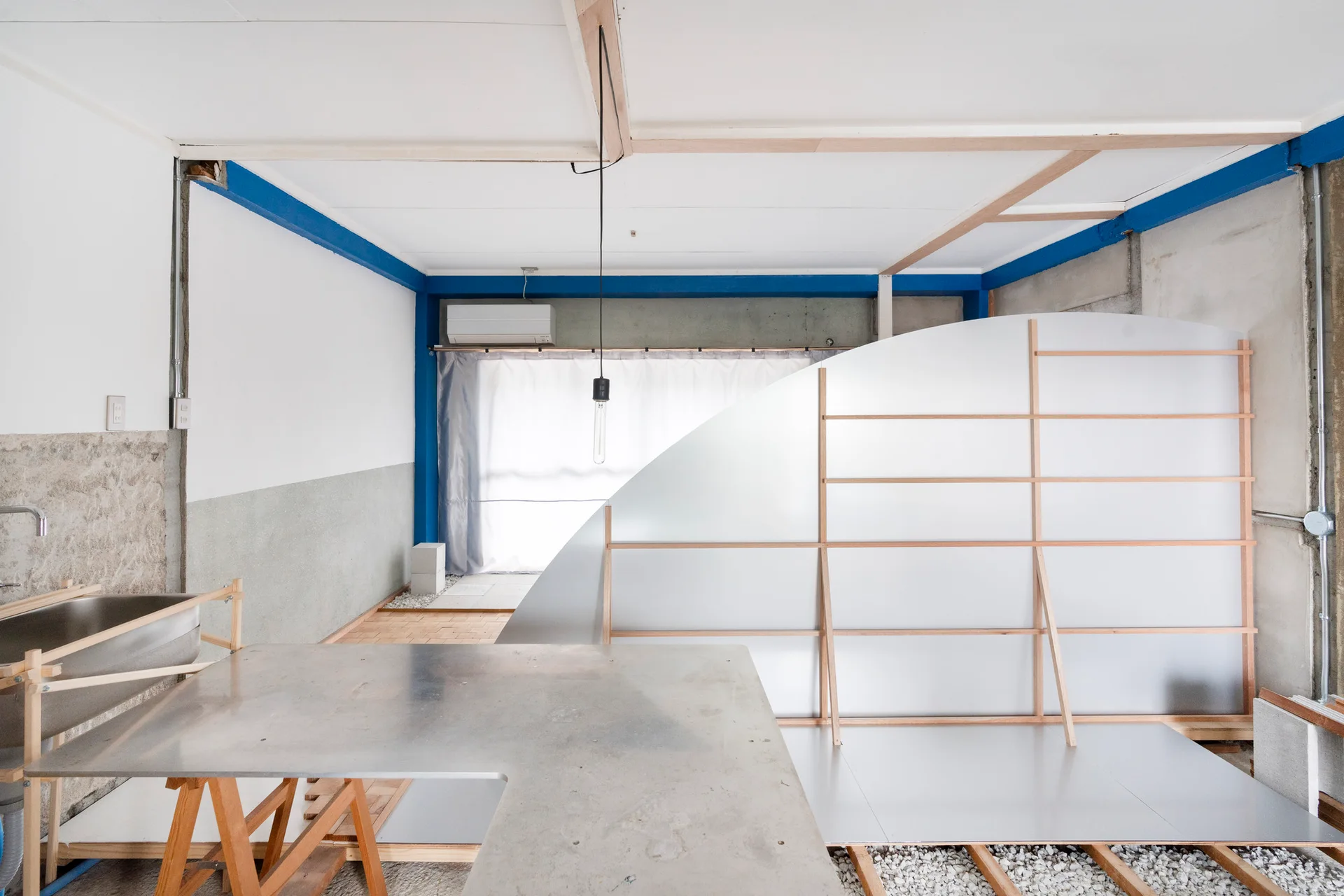 An industrial studio space features an L-shaped metal desk, a large curved partition with a wood frame, and blue painted ceiling beams.