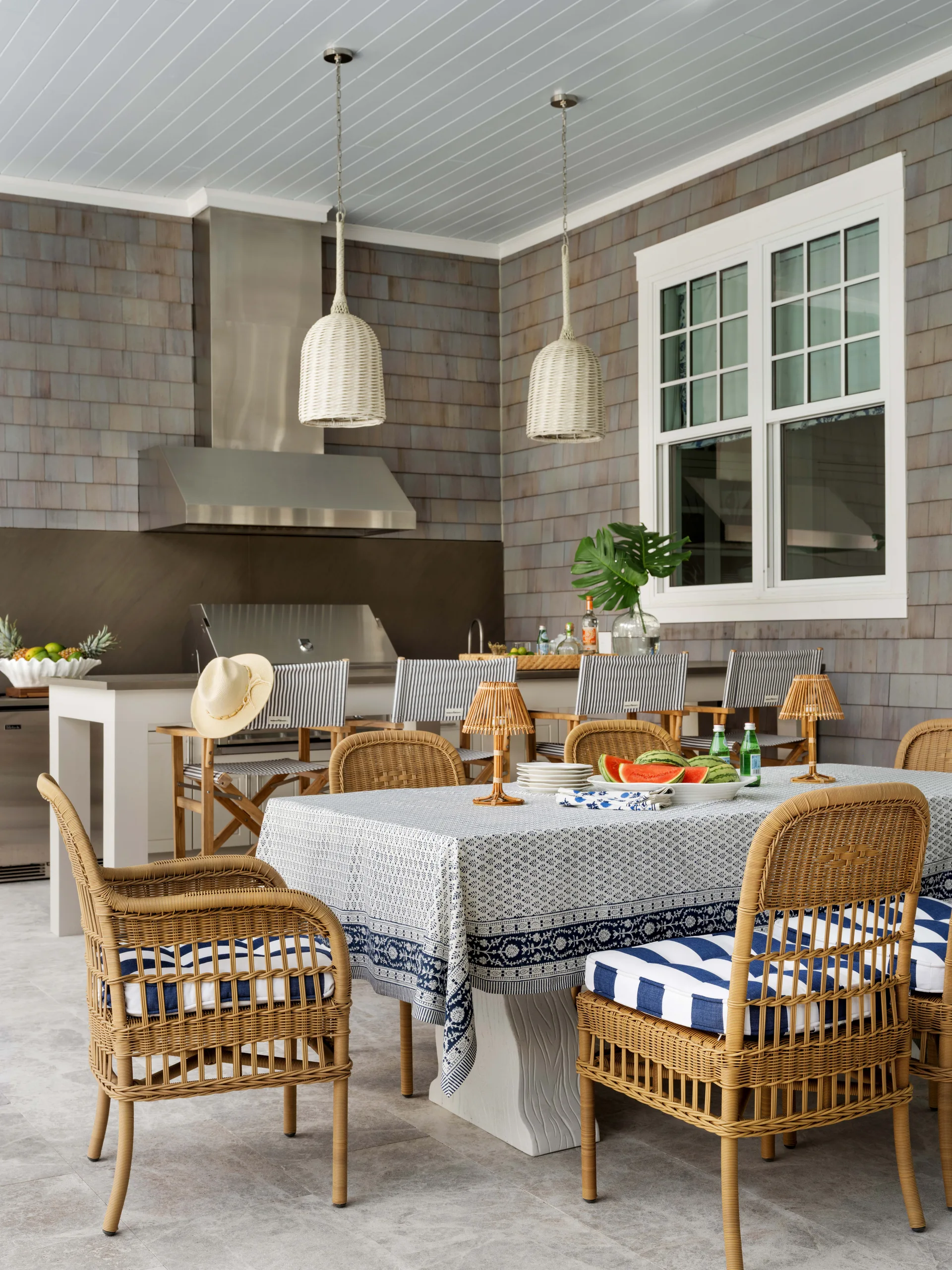 A covered outdoor patio features a dining table with blue and white linens, wicker chairs, and a stainless steel grill kitchen against a shingled wall.