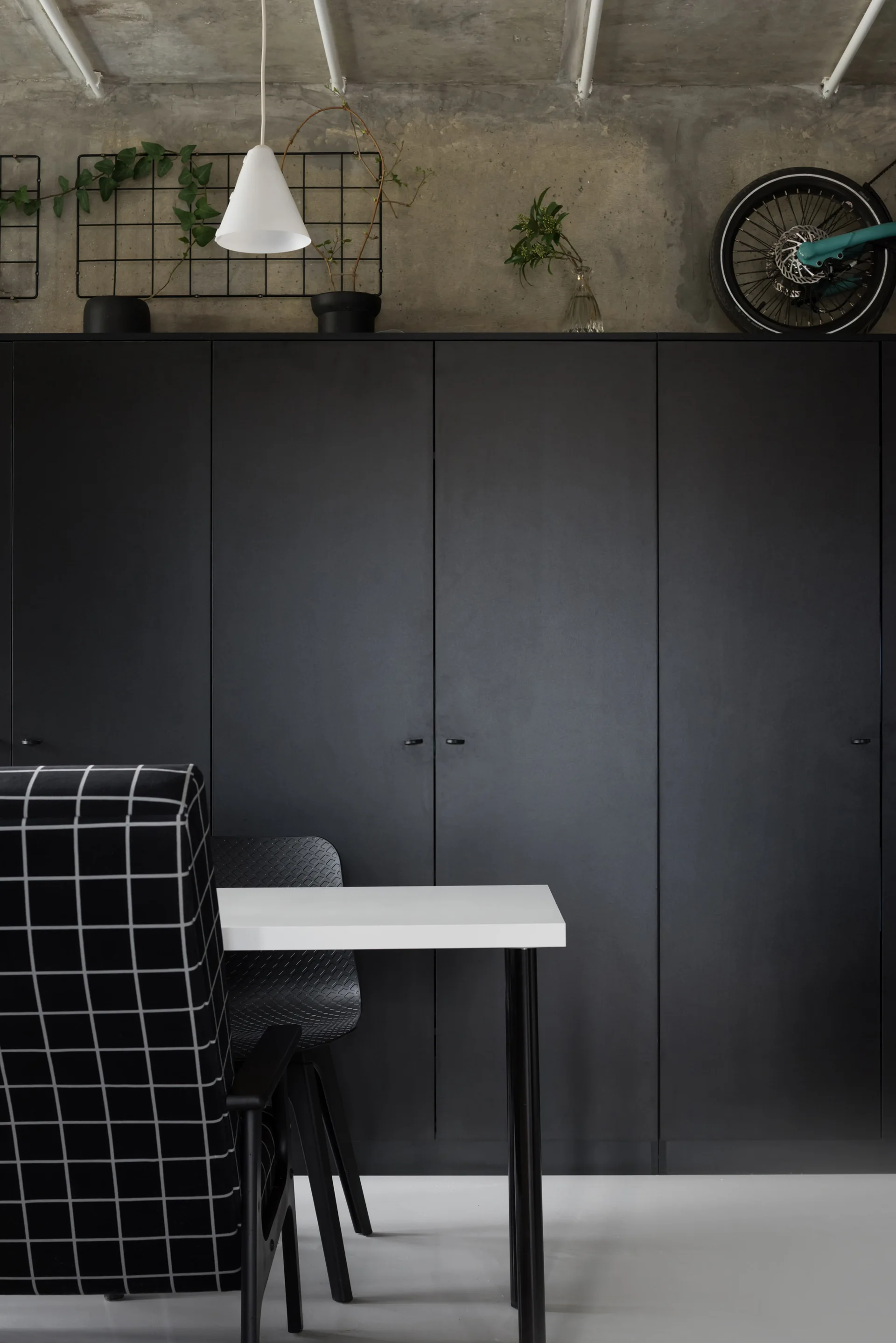 A small dining table and grid-patterned chair sit before a floor-to-ceiling matte black cabinet under an exposed concrete ceiling.