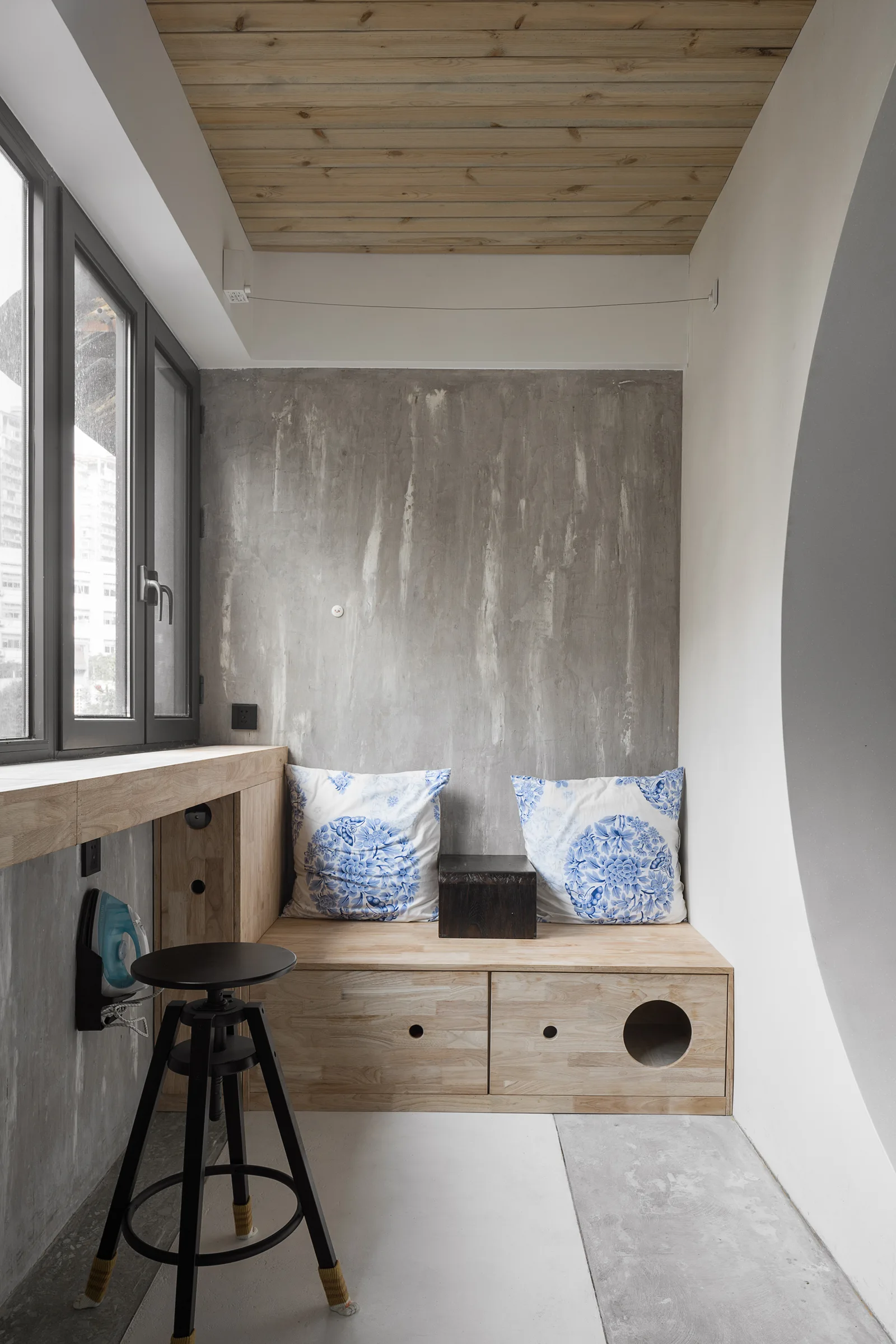 A small alcove features raw concrete walls, a light wood plank ceiling, and a built-in wooden storage bench with blue and white floral cushions.