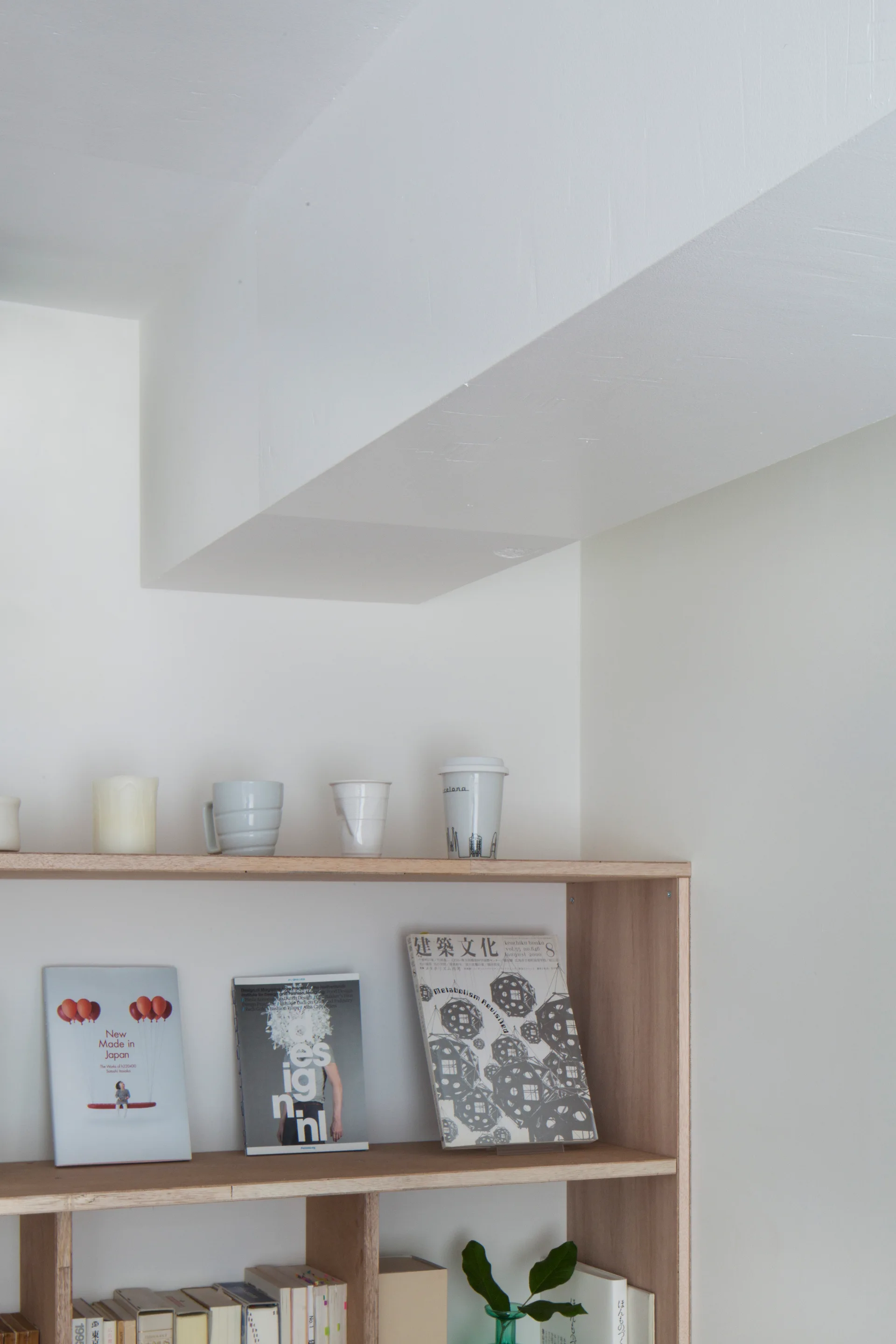 A minimalist light wood bookshelf styled with ceramic mugs and forward-facing books stands against a white wall beneath a ceiling soffit.