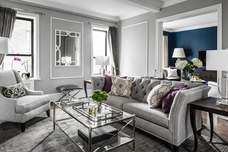 A grey living room features a tufted sofa, matching armchair, glass coffee table, and walls decorated with white molding panels.