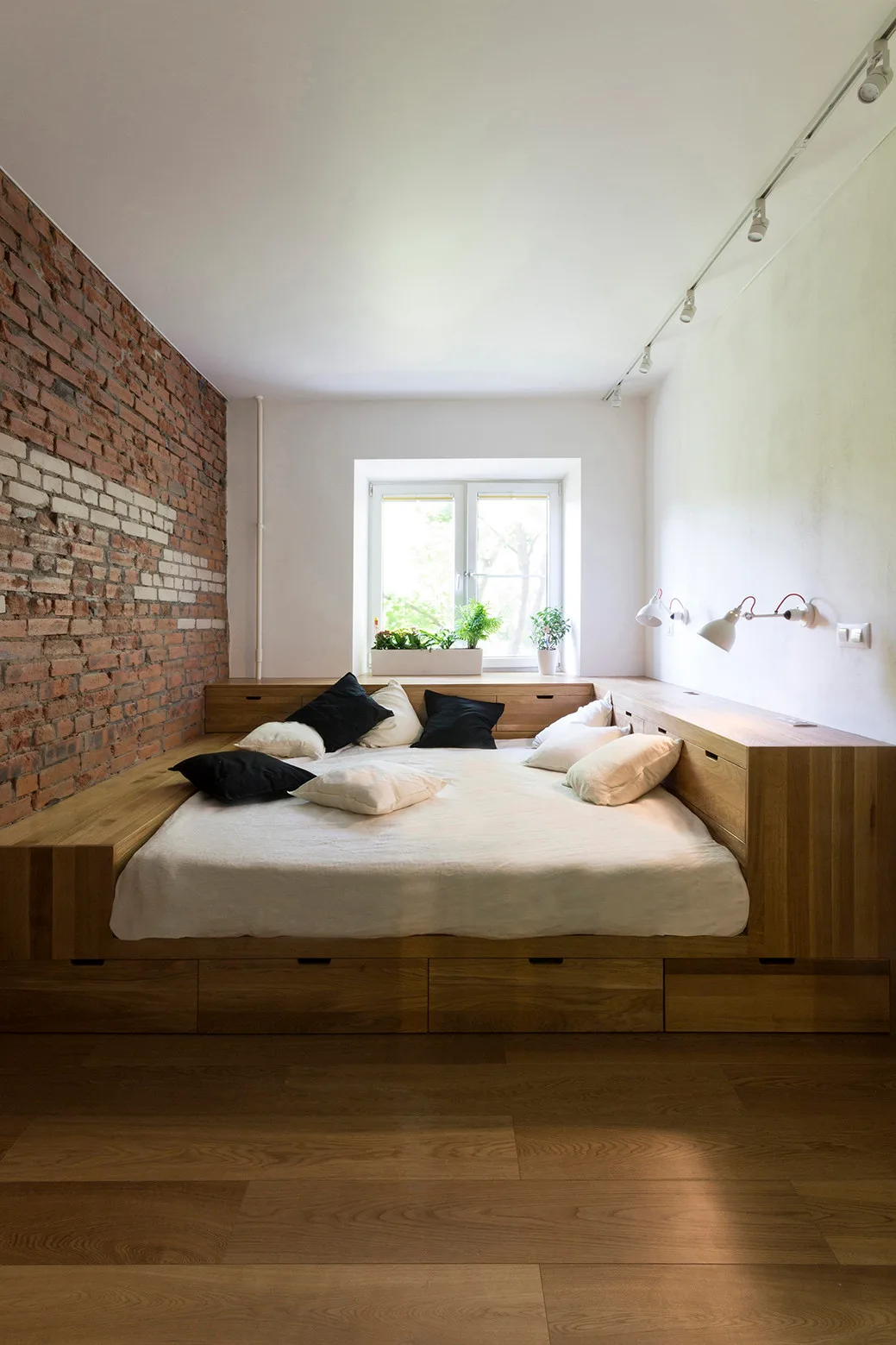 A custom low wooden platform bed with built-in drawers sits against an exposed red brick wall and white walls beneath a central window.