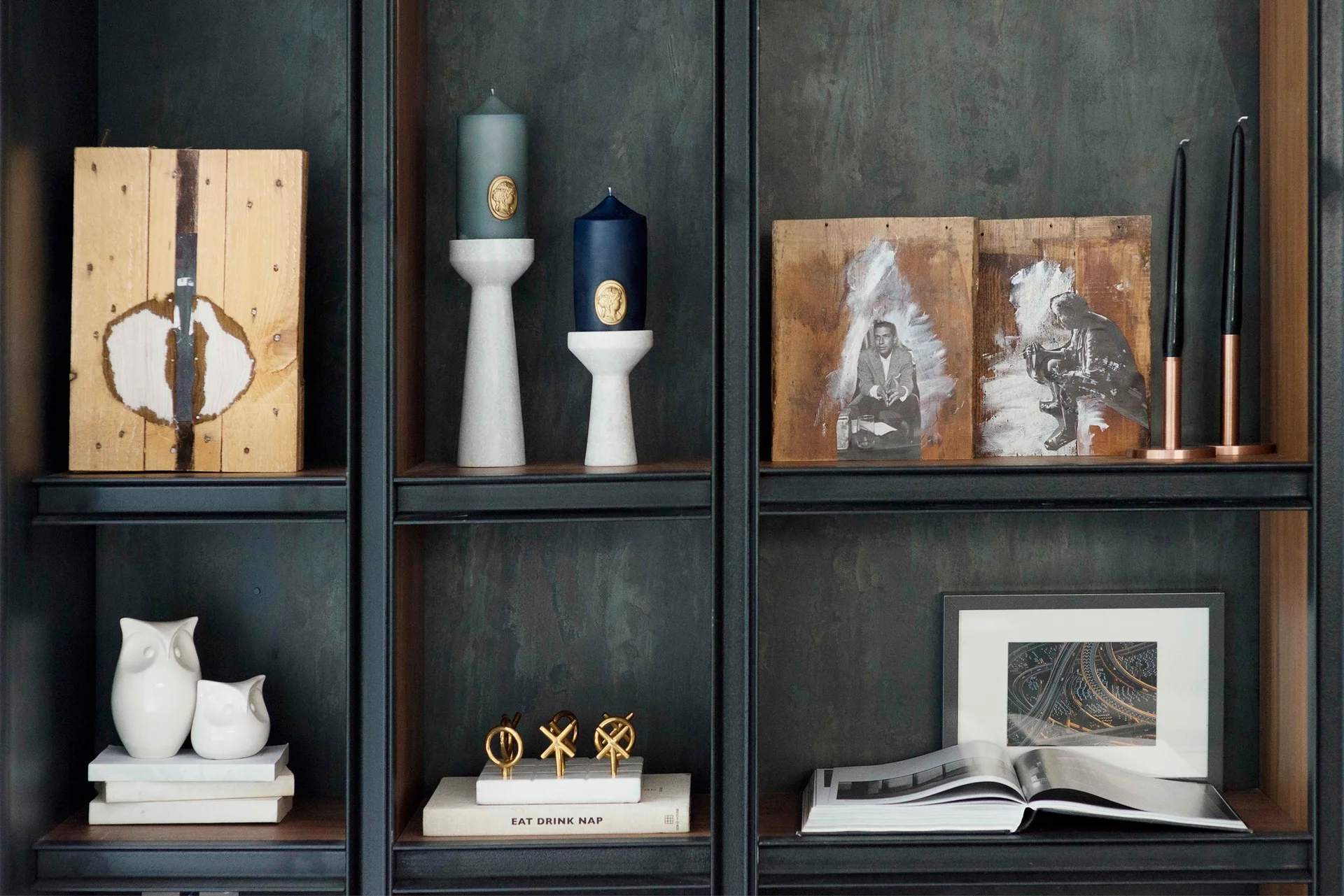 A dark charcoal metal open shelving unit styled with sculptural white candle holders, books, and wood-backed artwork.