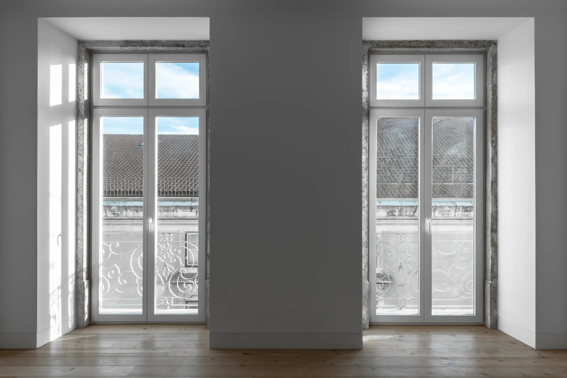 Two tall white windows framed in rough gray stone overlook a balcony in a room with light wood floors.