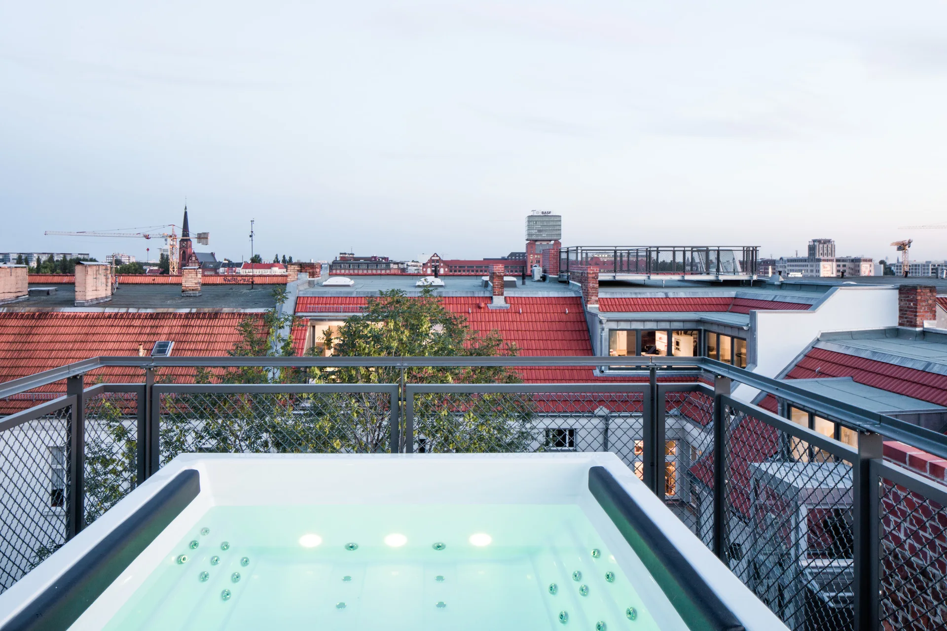 A glowing white hot tub sits on a rooftop terrace surrounded by a metal mesh railing, overlooking red-tiled city roofs at dusk.