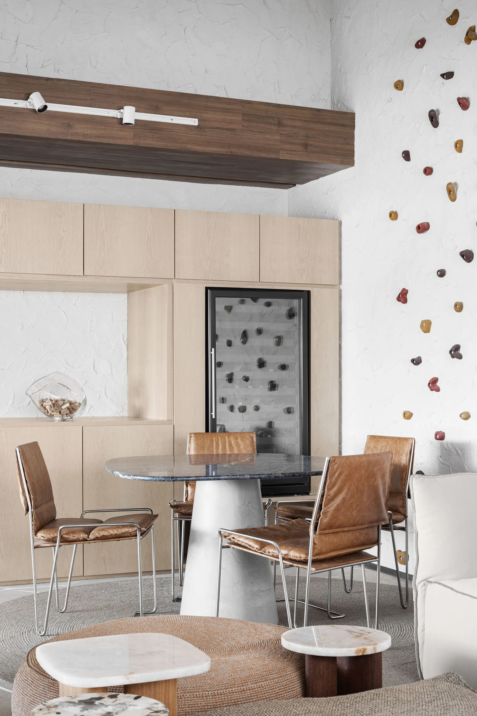 A round stone dining table and tan leather chairs are set against light wood cabinetry, with colorful climbing holds on the textured wall.