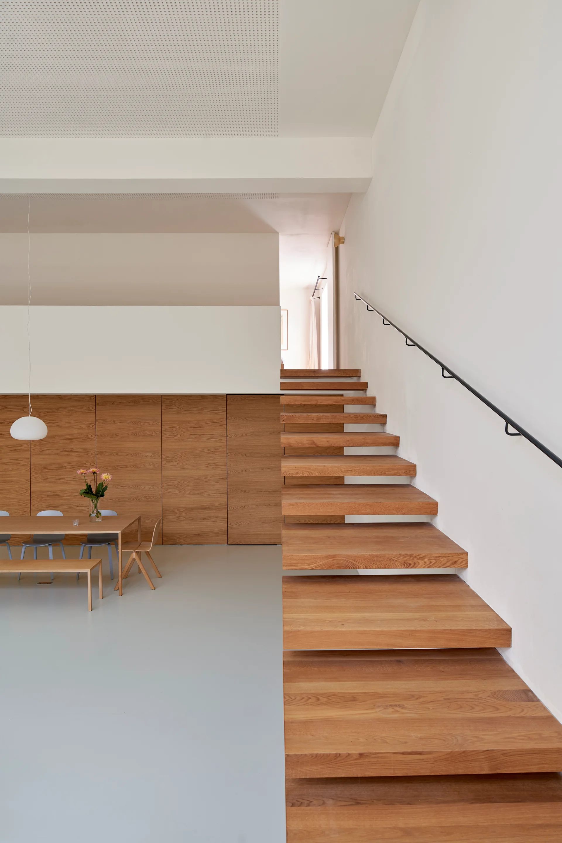 Floating wooden stairs with a black metal handrail lead up a white wall beside a dining table and wood paneled storage cabinets on grey flooring.