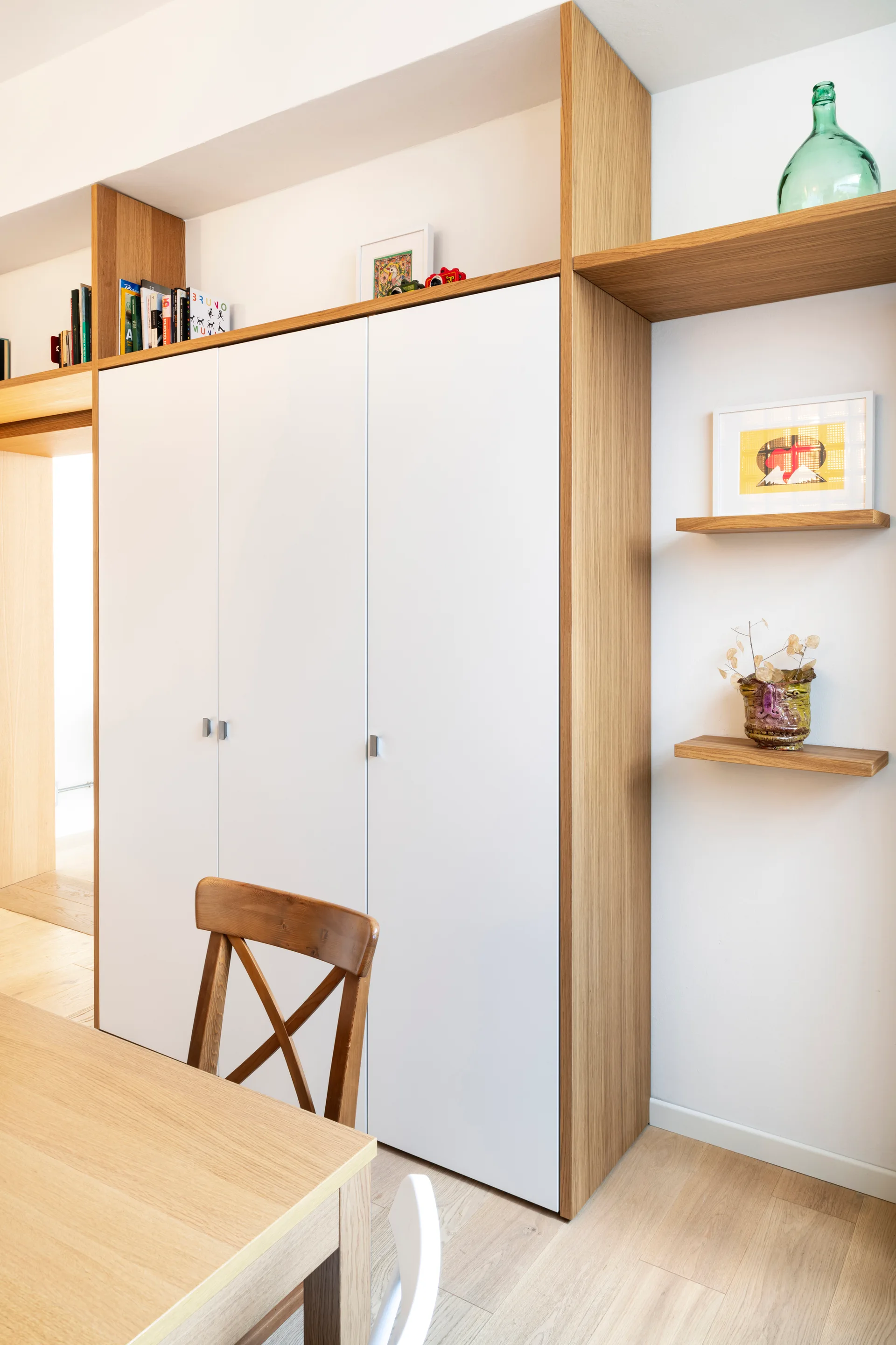 A custom storage unit with white flat-front doors and a light wood frame stands beside floating oak shelves and a wooden cross-back chair.