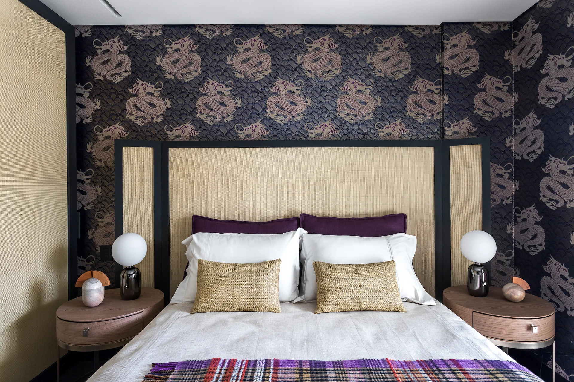 A symmetrical bedroom with a tan woven headboard against purple dragon-patterned wallpaper, flanked by oval nightstands with globe lamps.