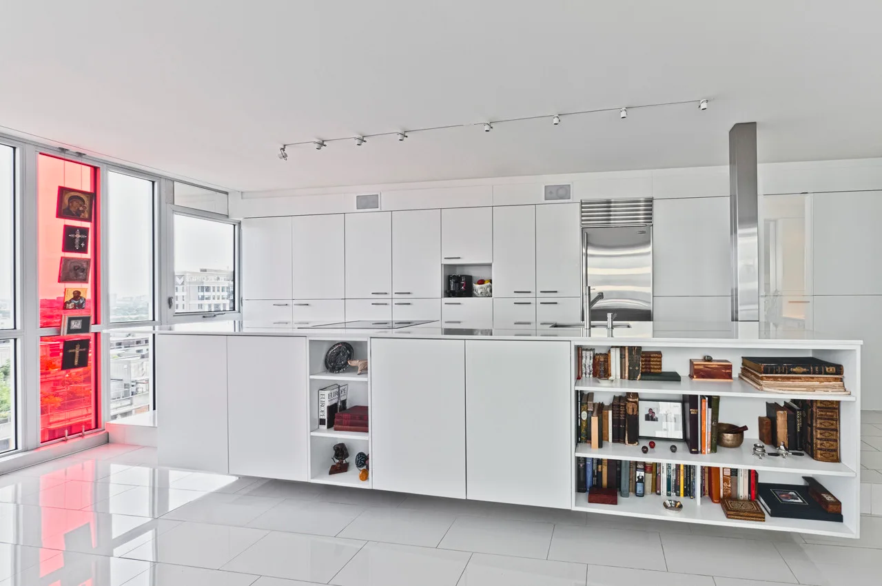 A minimalist white kitchen features floor-to-ceiling cabinets, a glossy tile floor, and a large central island with integrated open bookshelves.