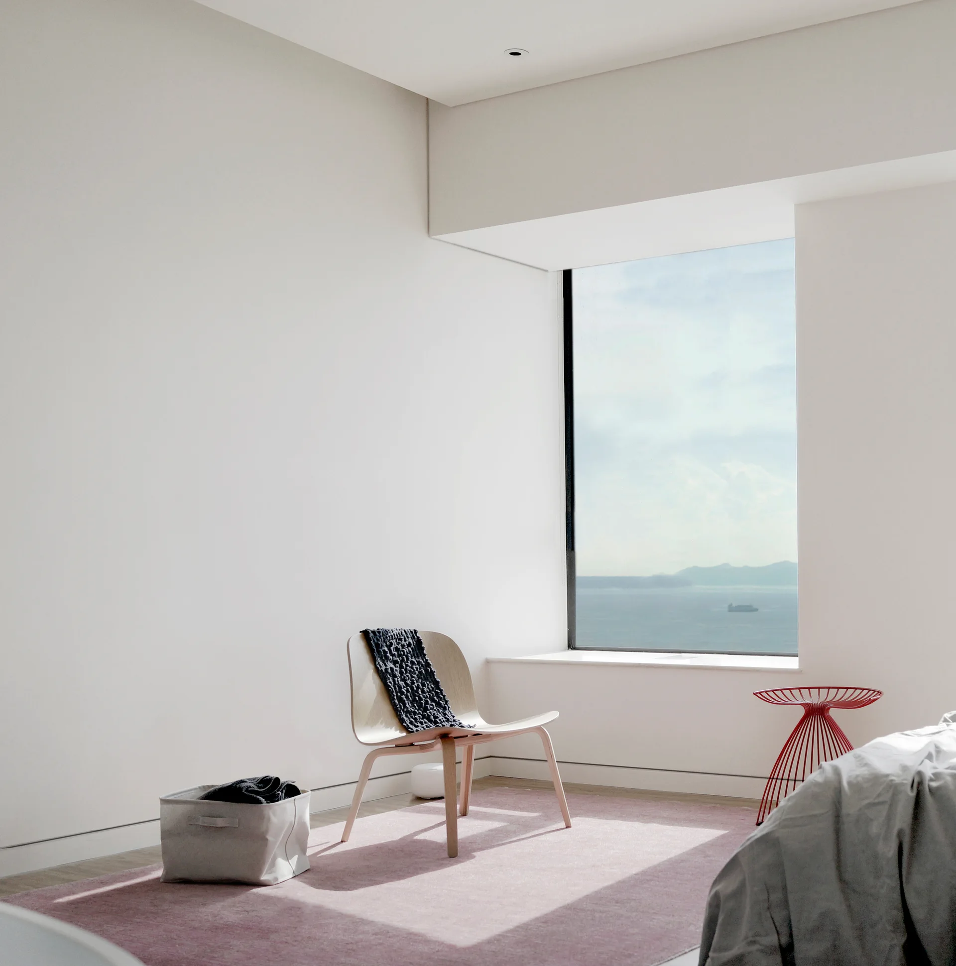 A minimalist bedroom featuring a curved wooden chair on a pink area rug next to a window overlooking the sea.