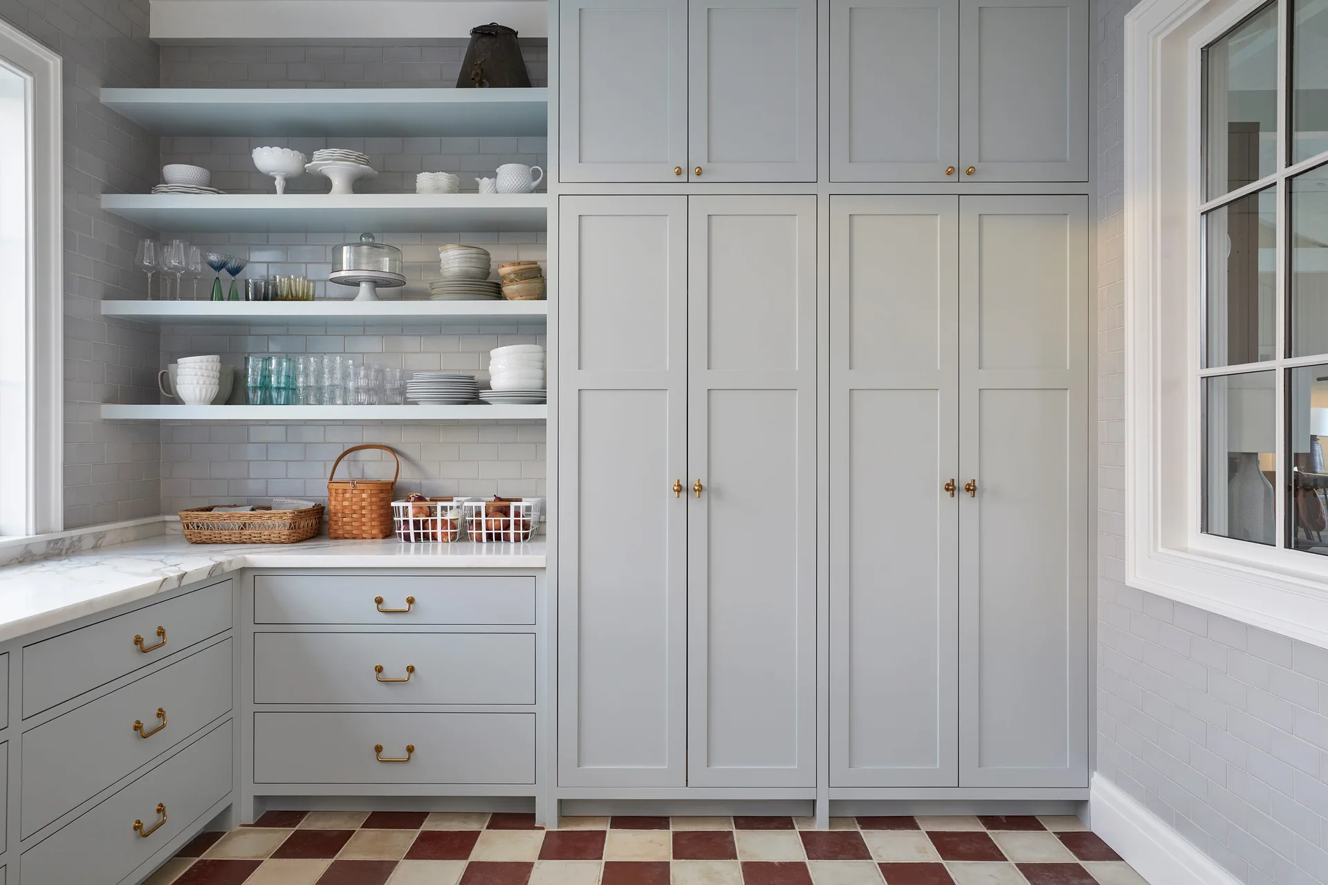 Light gray built-in pantry cabinets and open shelves on a matching tiled wall stand above a red and cream checkerboard tile floor.