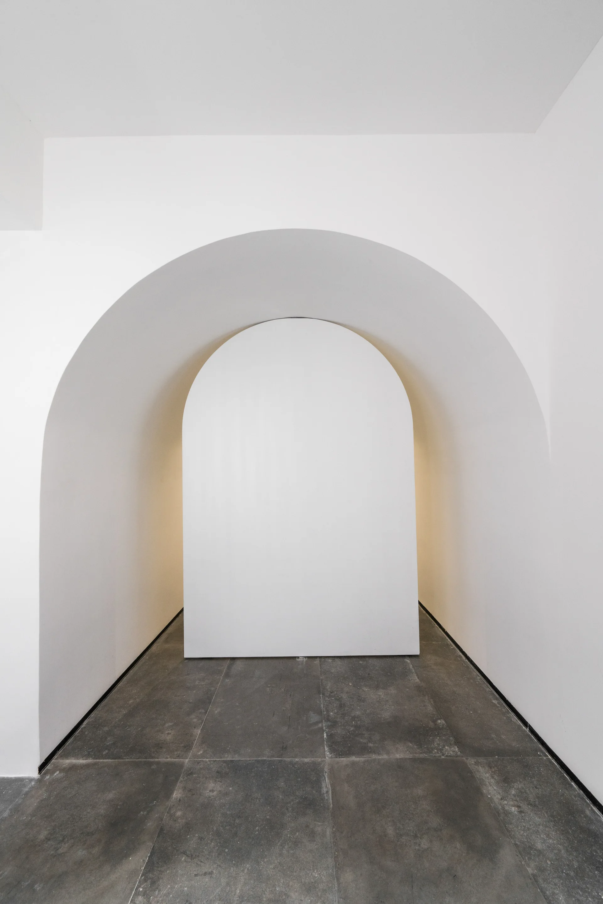 A minimalist hallway features a white arched partition wall centered inside a larger archway, set over dark grey stone floor tiles.